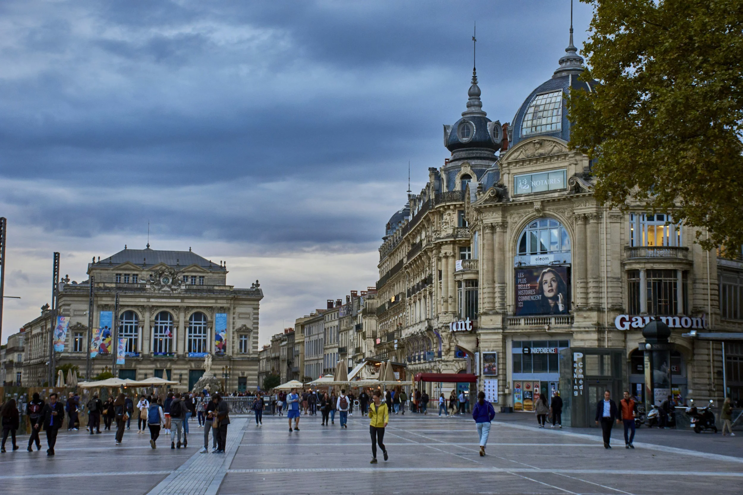 A busy city square with historic buildings, outdoor cafes, and many pedestrians under a cloudy sky.