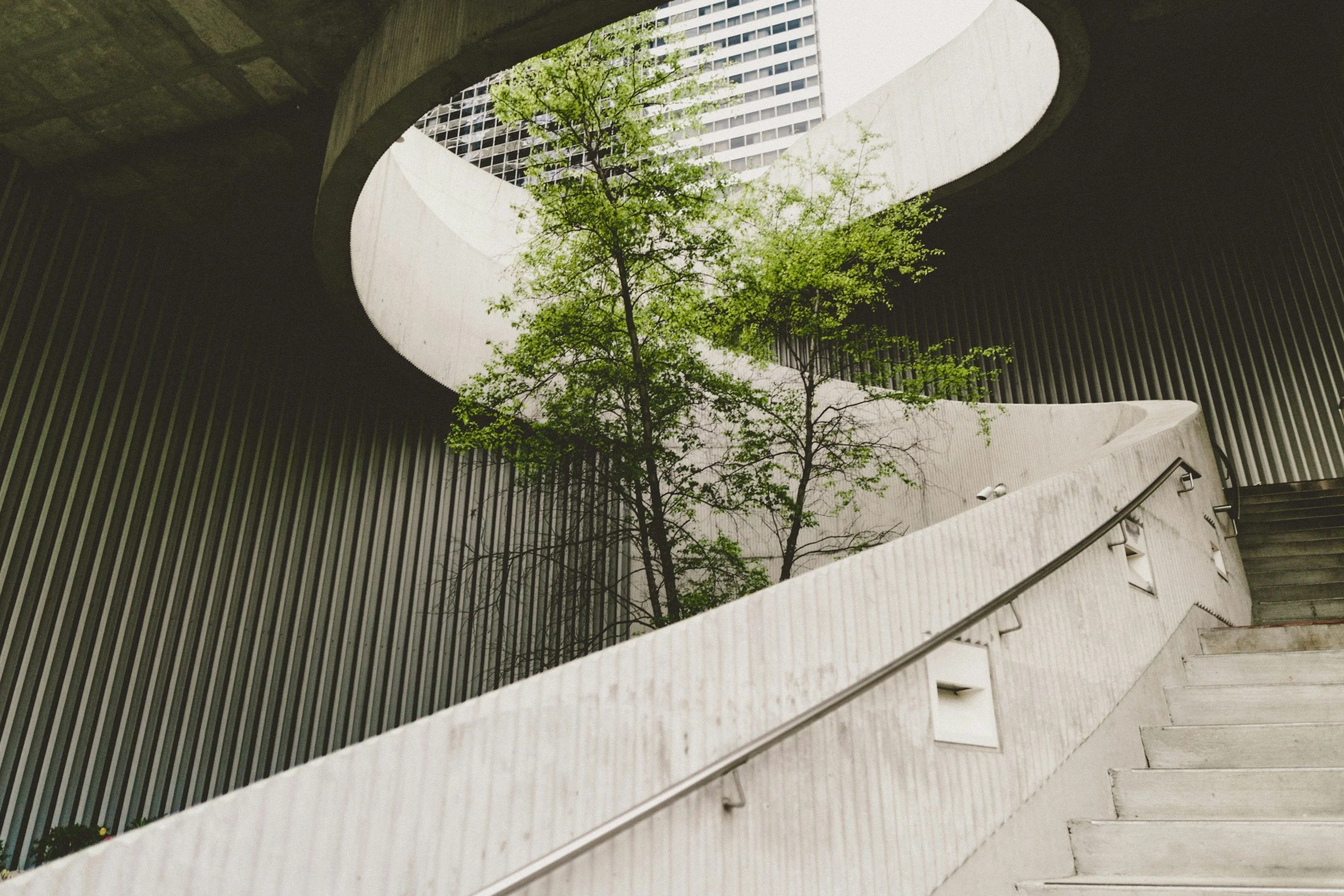 Looking up from the bottom of an interior spiral staircase, stairs made of concrete with metal railing, the staircase surrounds a courtyard with green trees, surrounded by modern building with glass windows.