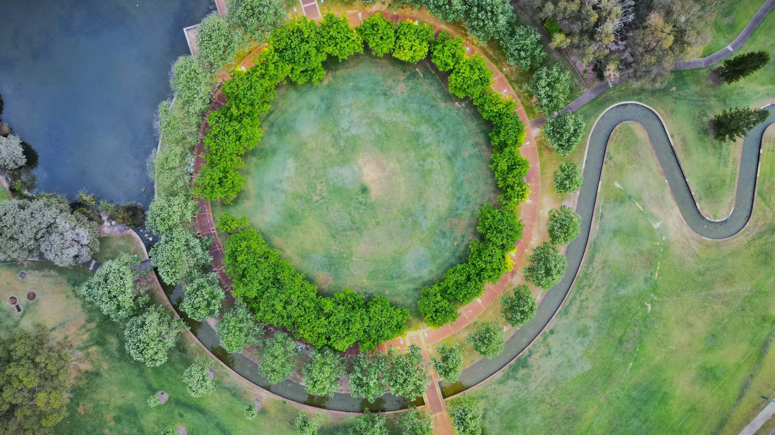 An aerial view of a park with a circular grassy area surrounded by trees, a water body on the left, and winding pathways on the right.