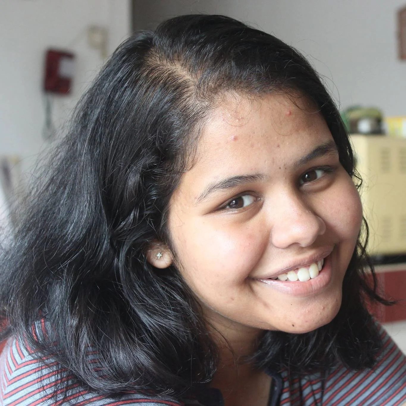 Close-up of a young woman with dark, wavy hair and a bright, smiling face, wearing a striped shirt and a small earring.