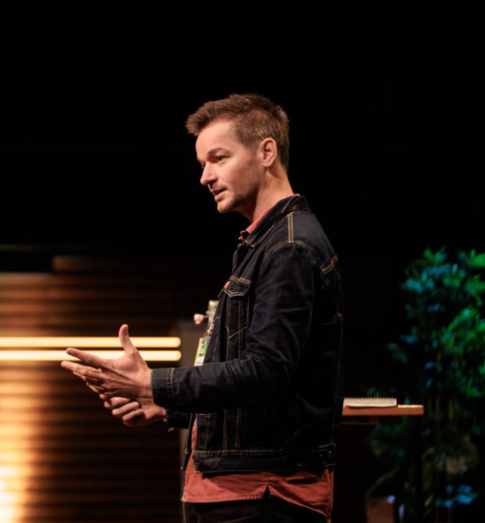 A man in a denim jacket speaking or presenting on stage, with a dark background and some green plants visible.