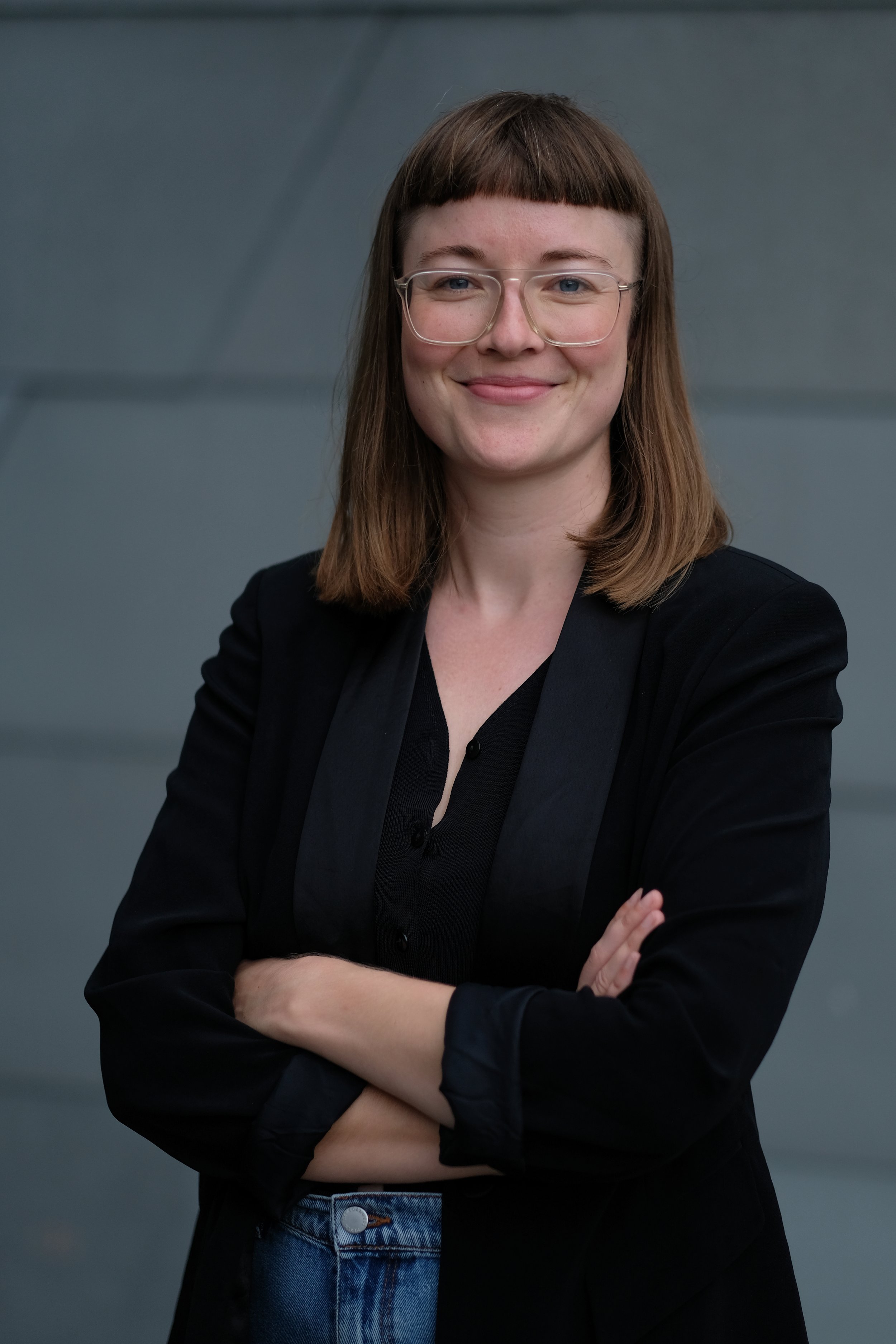 A woman with shoulder-length brown hair, wearing glasses, a black blazer, black shirt, and blue jeans, standing with crossed arms against a dark gray background.