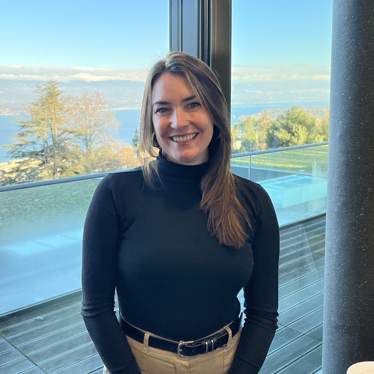 A smiling woman in a black turtleneck sweater standing indoors near a large window with a view of trees, water, and a blue sky with clouds.