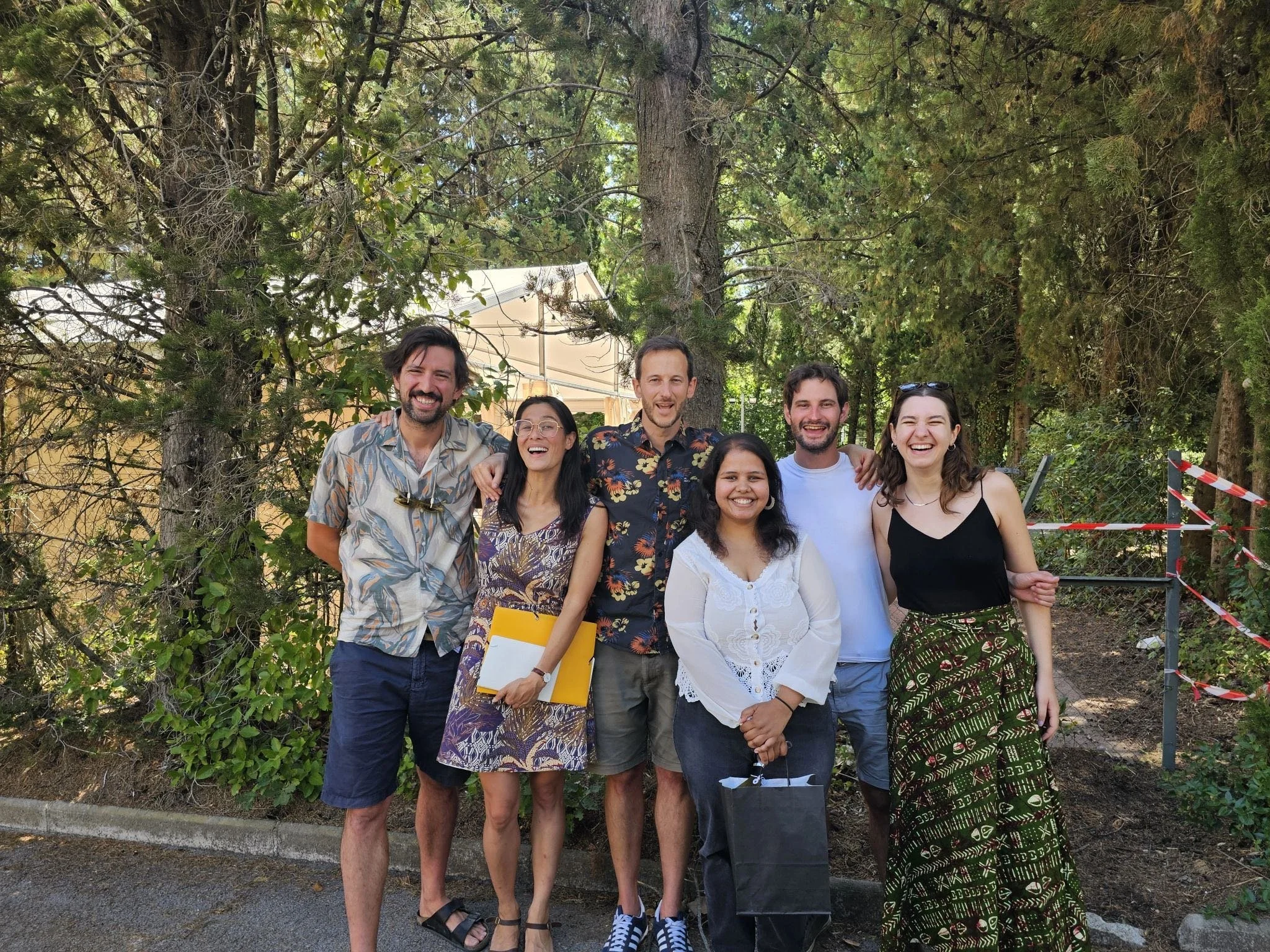 A group of seven diverse friends smiling and standing outdoors in front of trees, with an outdoor tent and red-and-white striped tape in the background.