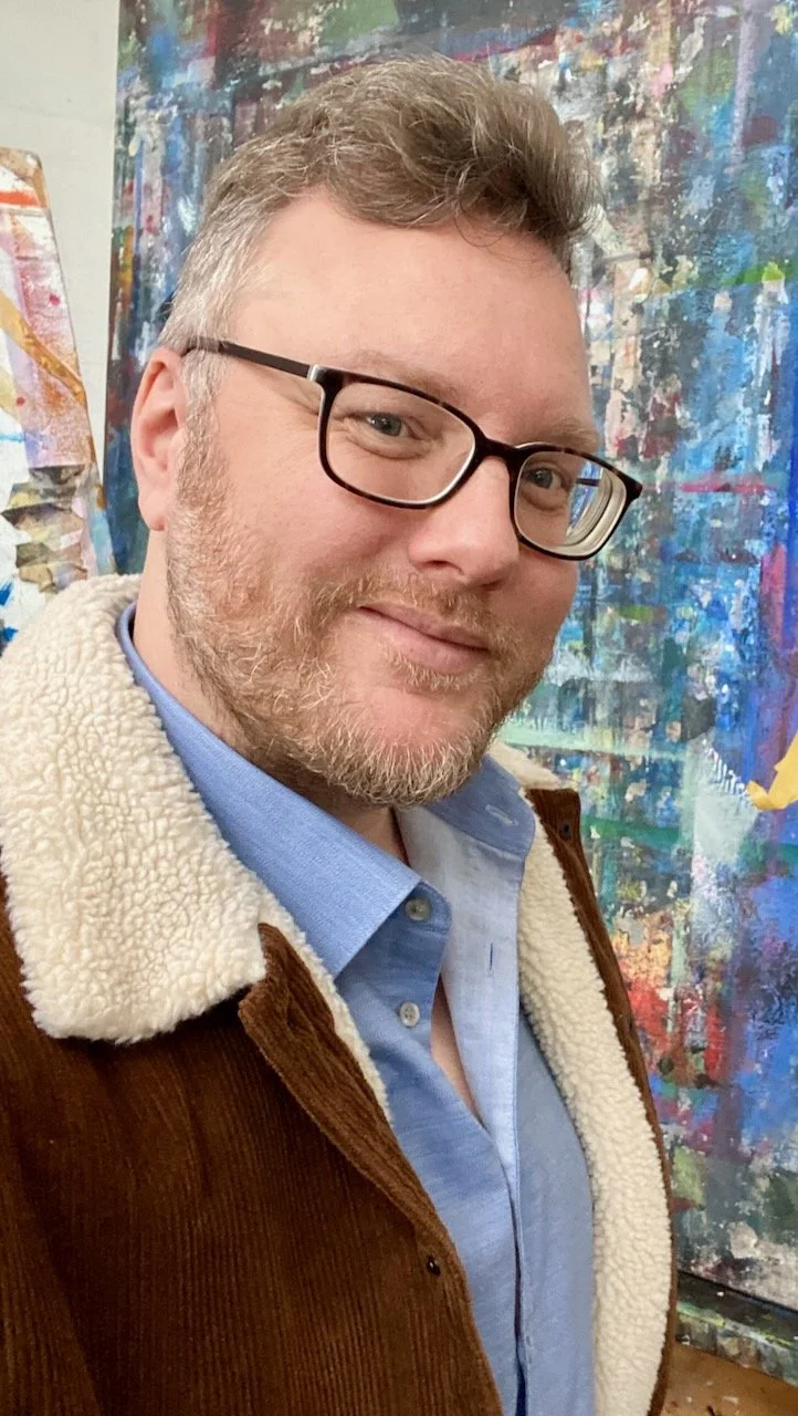 A man with glasses and a beard smiling for a selfie in front of a colorful abstract painting.