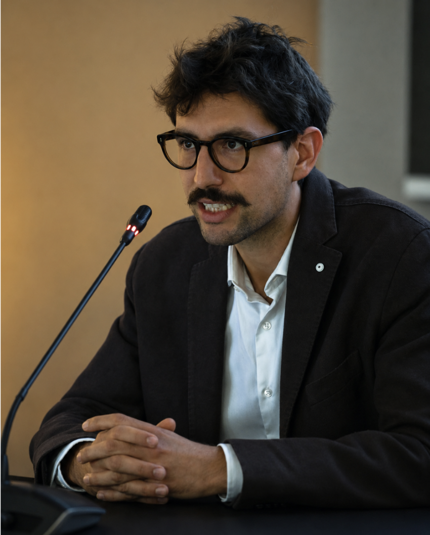 A man with dark curly hair, glasses, and a mustache is speaking into a microphone during a meeting or conference.