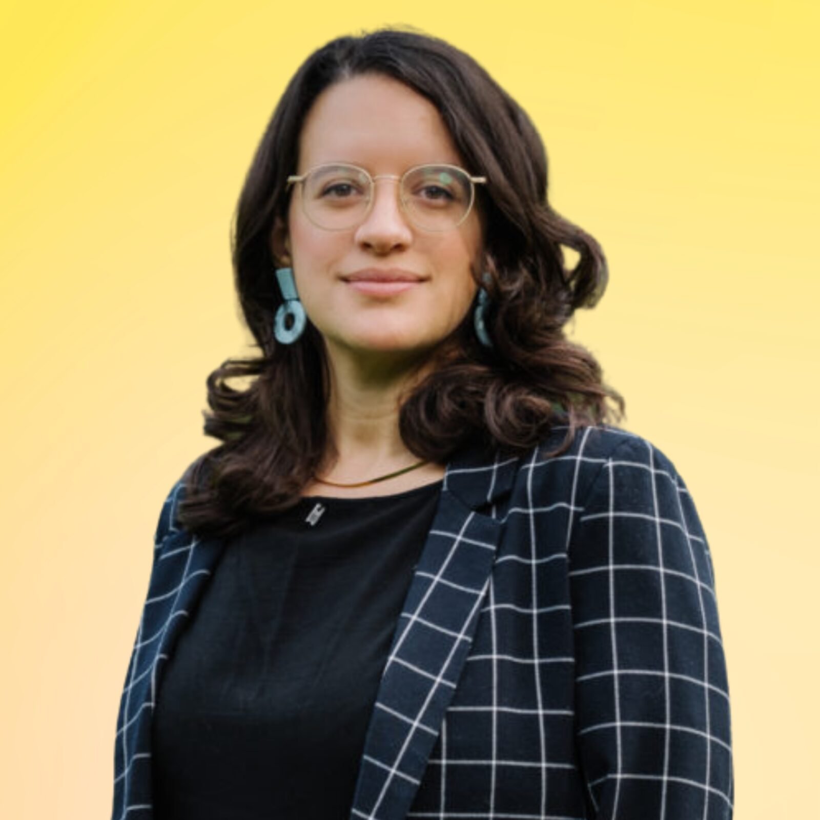 A woman with shoulder-length wavy dark hair, wearing glasses, earrings, a black top, and a blue checked blazer, standing against a yellow background.