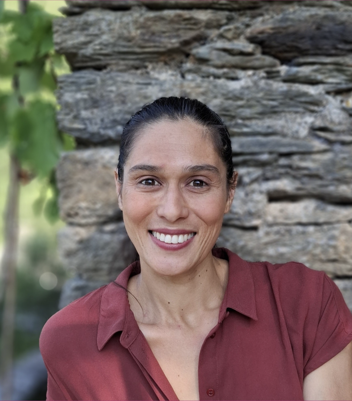 A woman smiling outdoors with a stone wall and greenery in the background.
