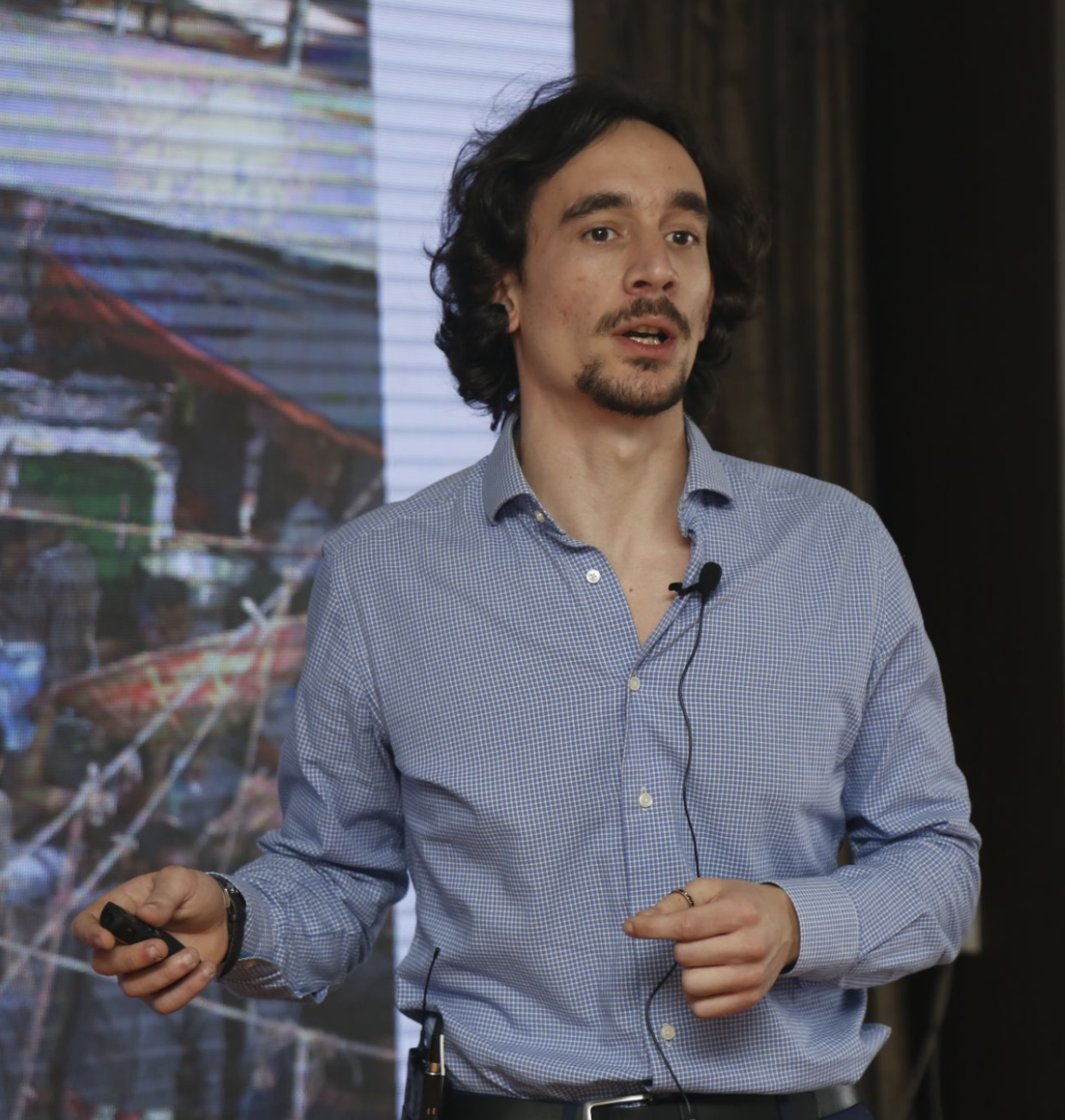 A man with shoulder-length dark hair, a beard, and wearing a blue checkered shirt is giving a presentation. He has a microphone clipped to his shirt and is holding a remote control in his right hand. The background shows a large screen or digital display with colorful images of a marketplace or outdoor scene.