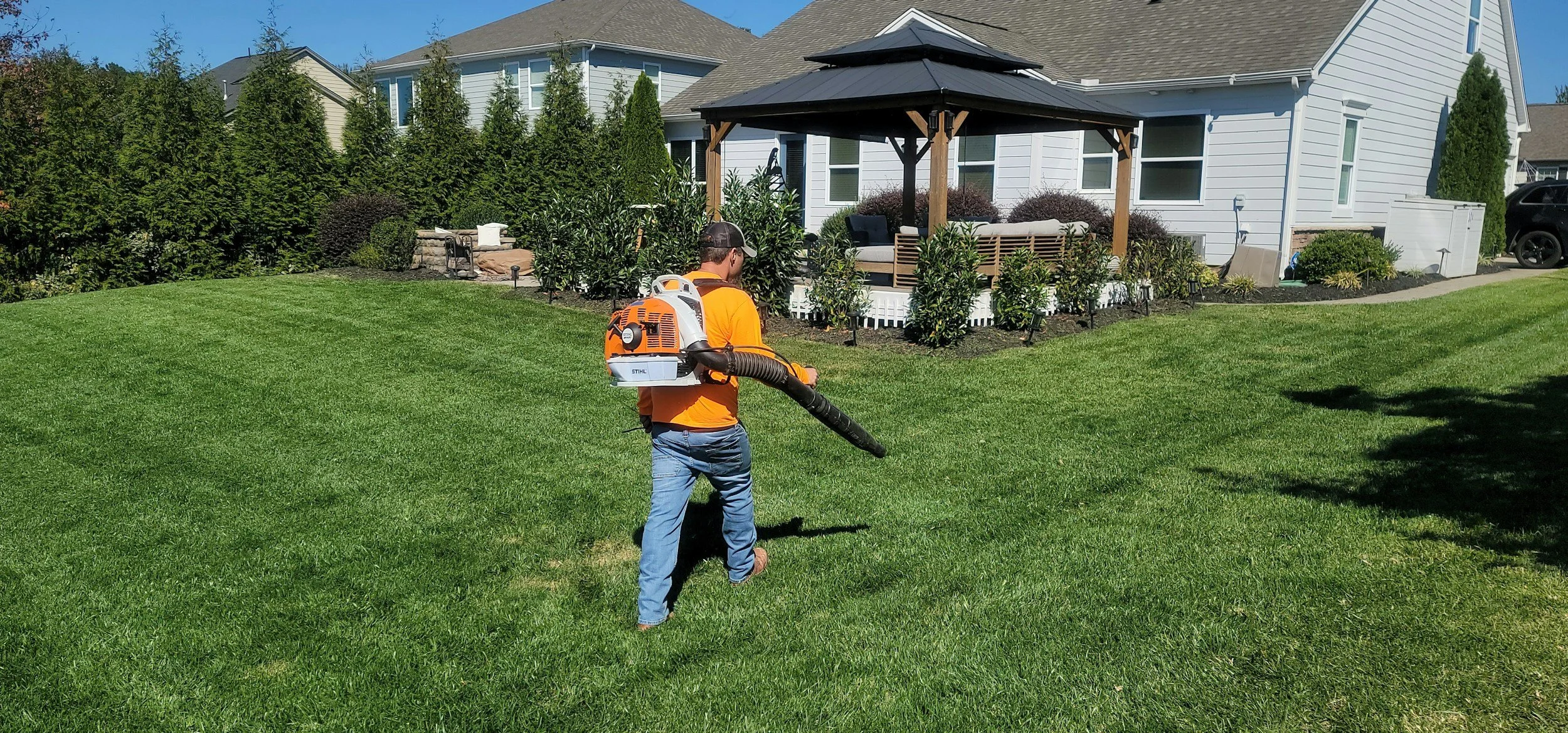 A man in an orange shirt and blue jeans is using a leaf blower on a green lawn in a backyard. In the background, there is a white house with a deck, patio furniture, and bushes.