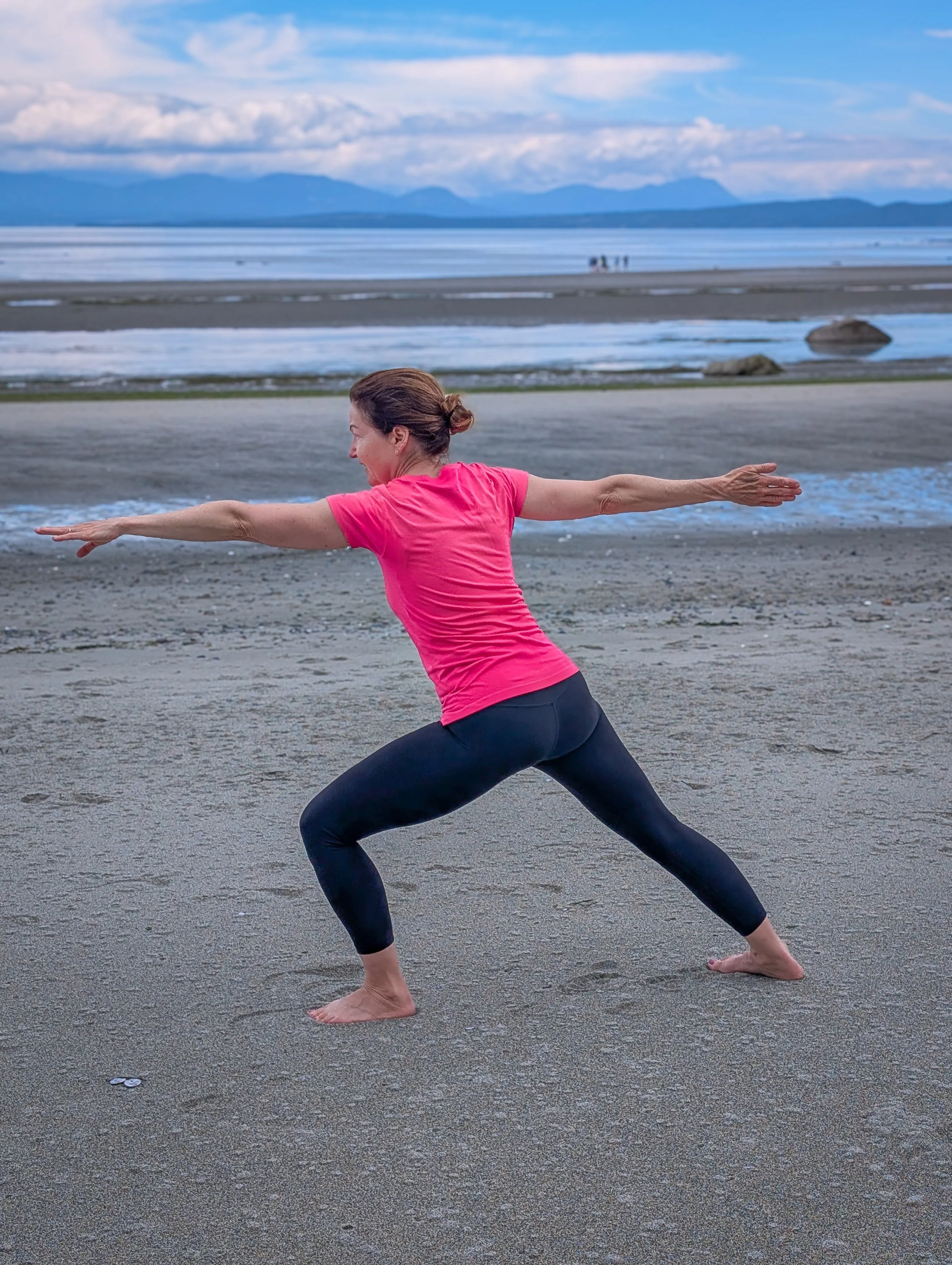 A woman practicing yoga on a beach near the water, wearing a pink shirt and black leggings, in a wide stance with arms extended.