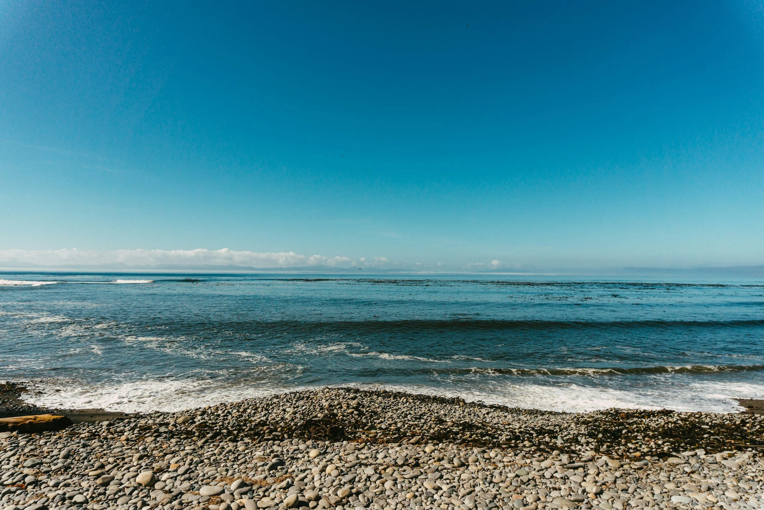 A rocky beach with small stones, calm ocean waves, and a clear blue sky with some clouds on the horizon.