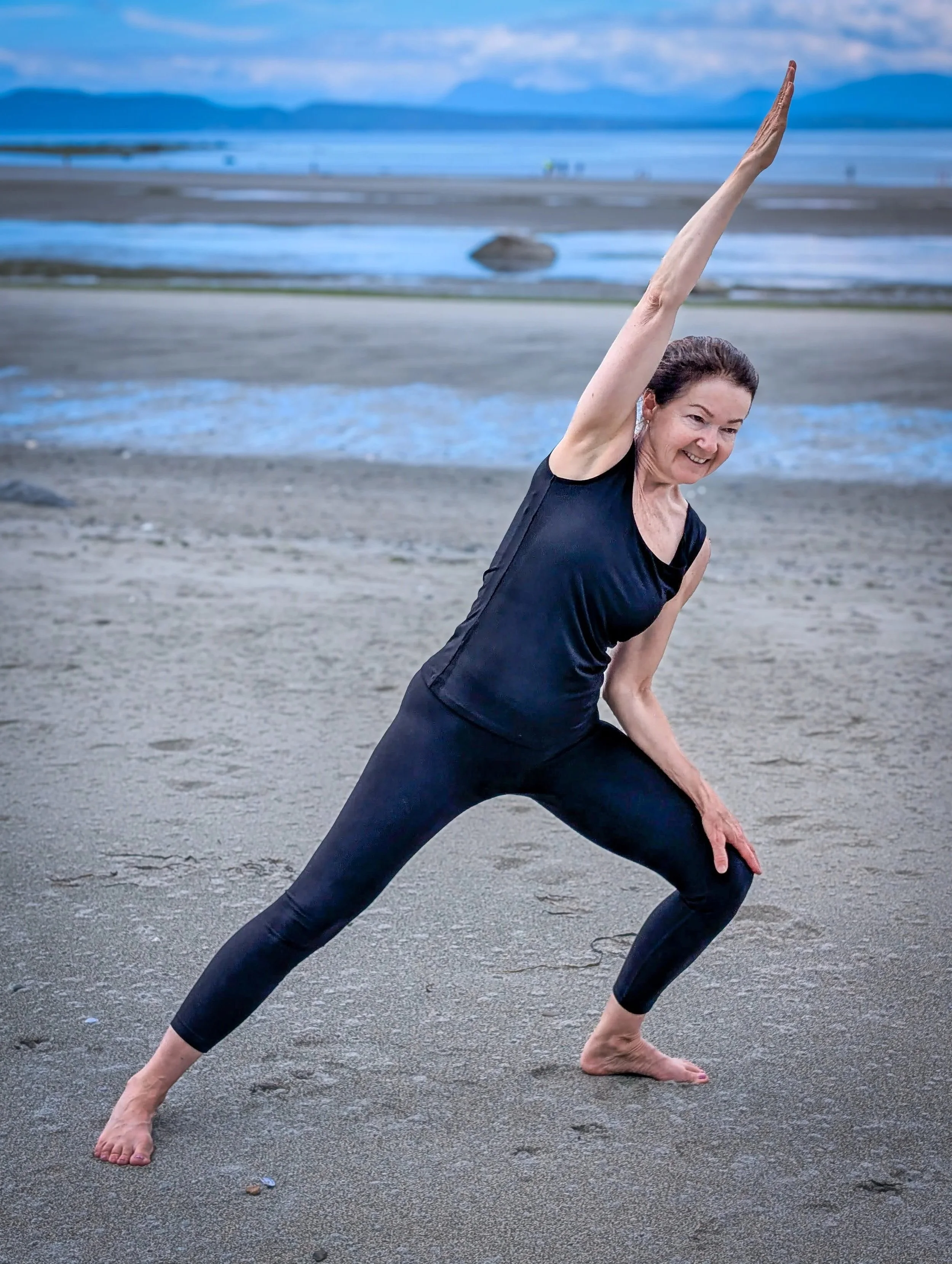 A woman practicing yoga on a beach, performing a wide-legged forward bend stretch. She is wearing a black tank top and black leggings, with an ocean and mountains in the background.