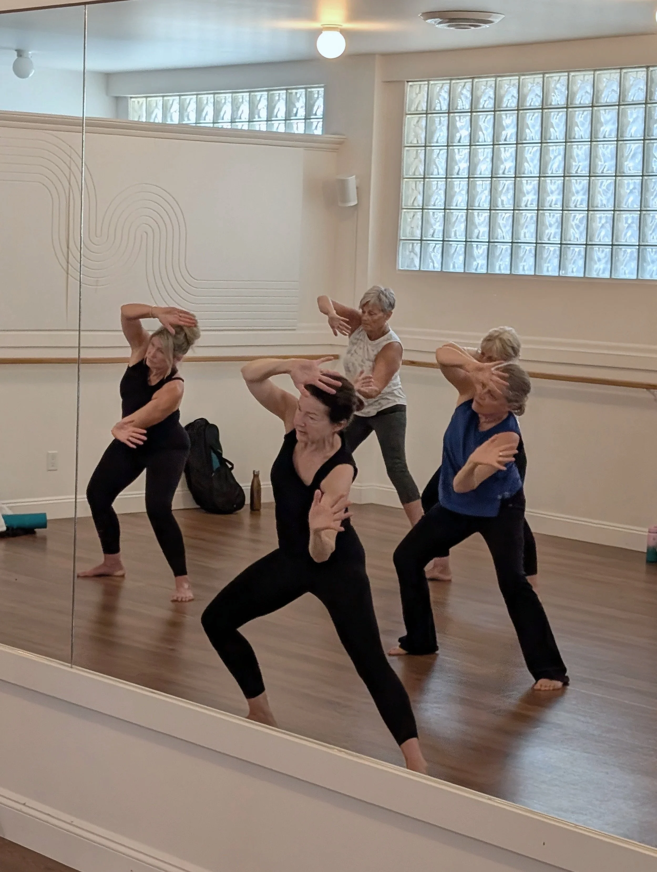 Group of people practicing yoga in a dance studio with mirrored wall and glass block windows.