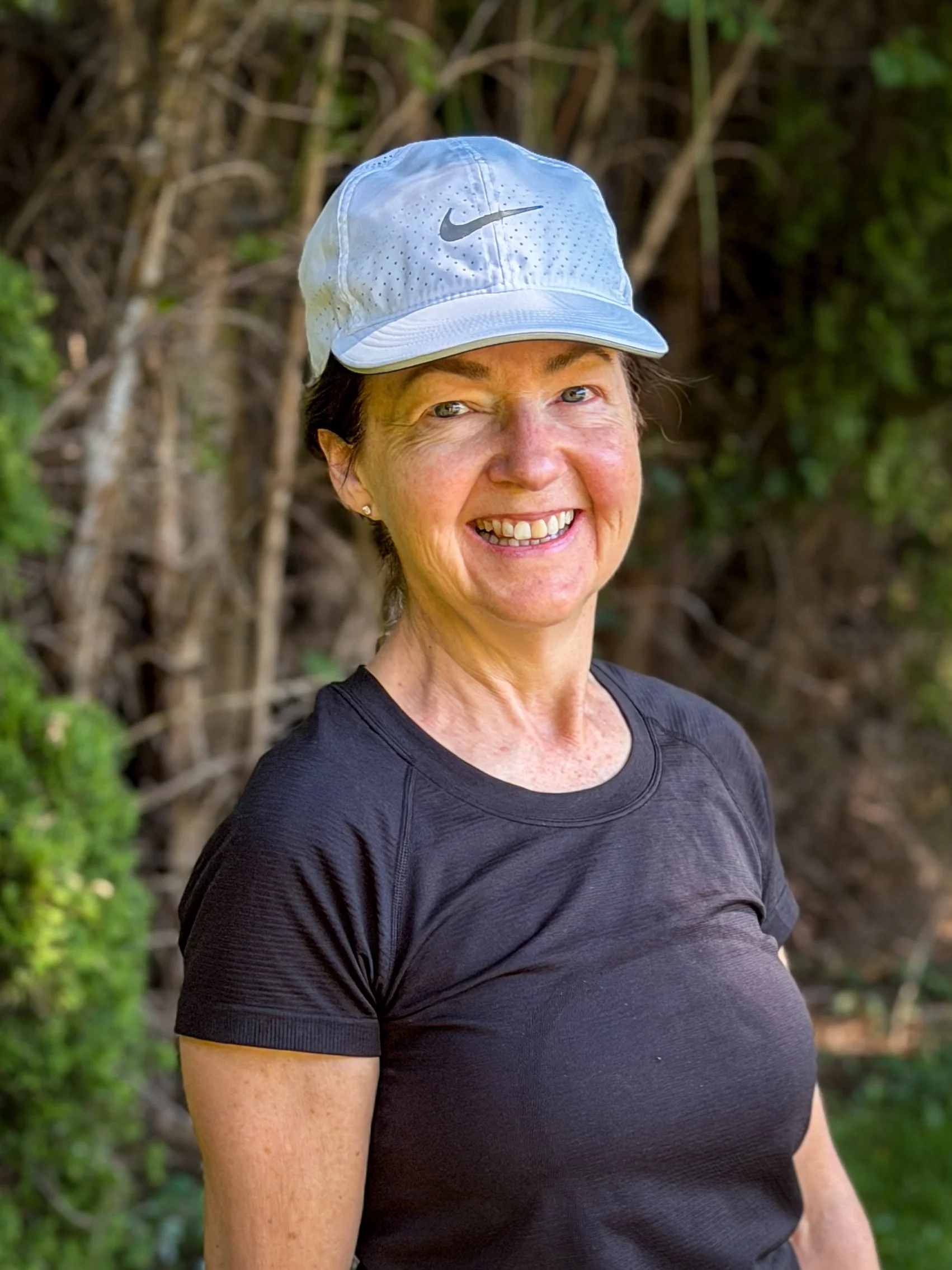 A woman smiling wearing a white Nike cap and a black athletic shirt outdoors surrounded by greenery.