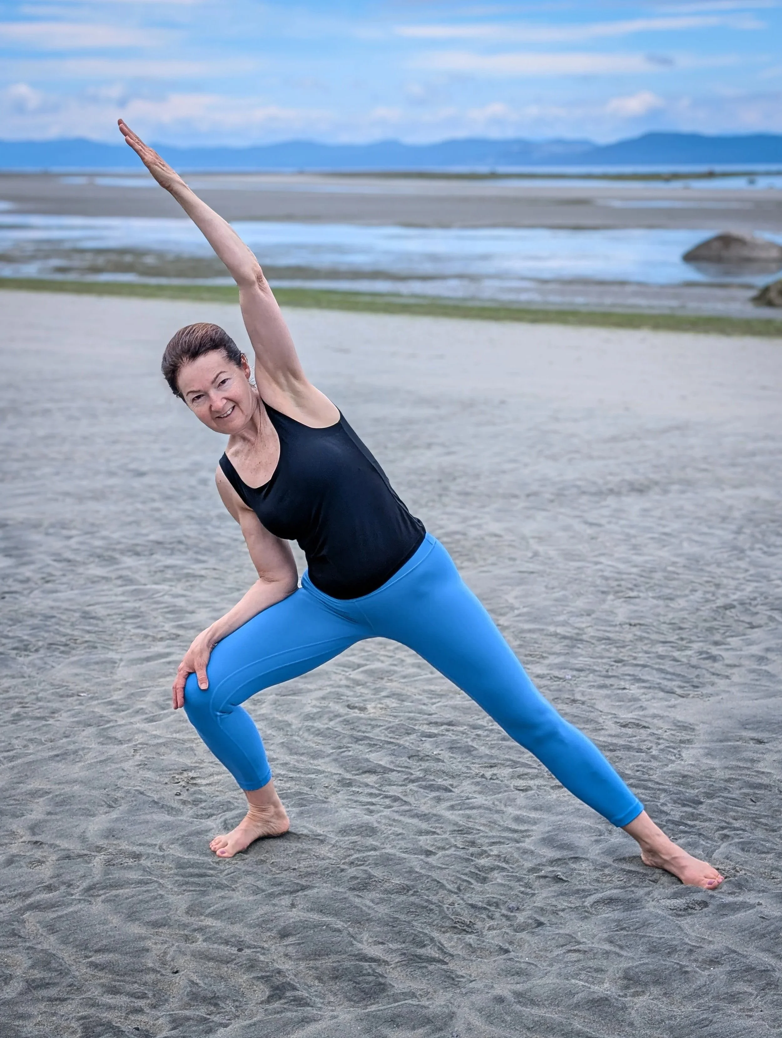 A woman practicing yoga on a sandy beach with mountains and water in the background, wearing a black tank top and blue leggings.