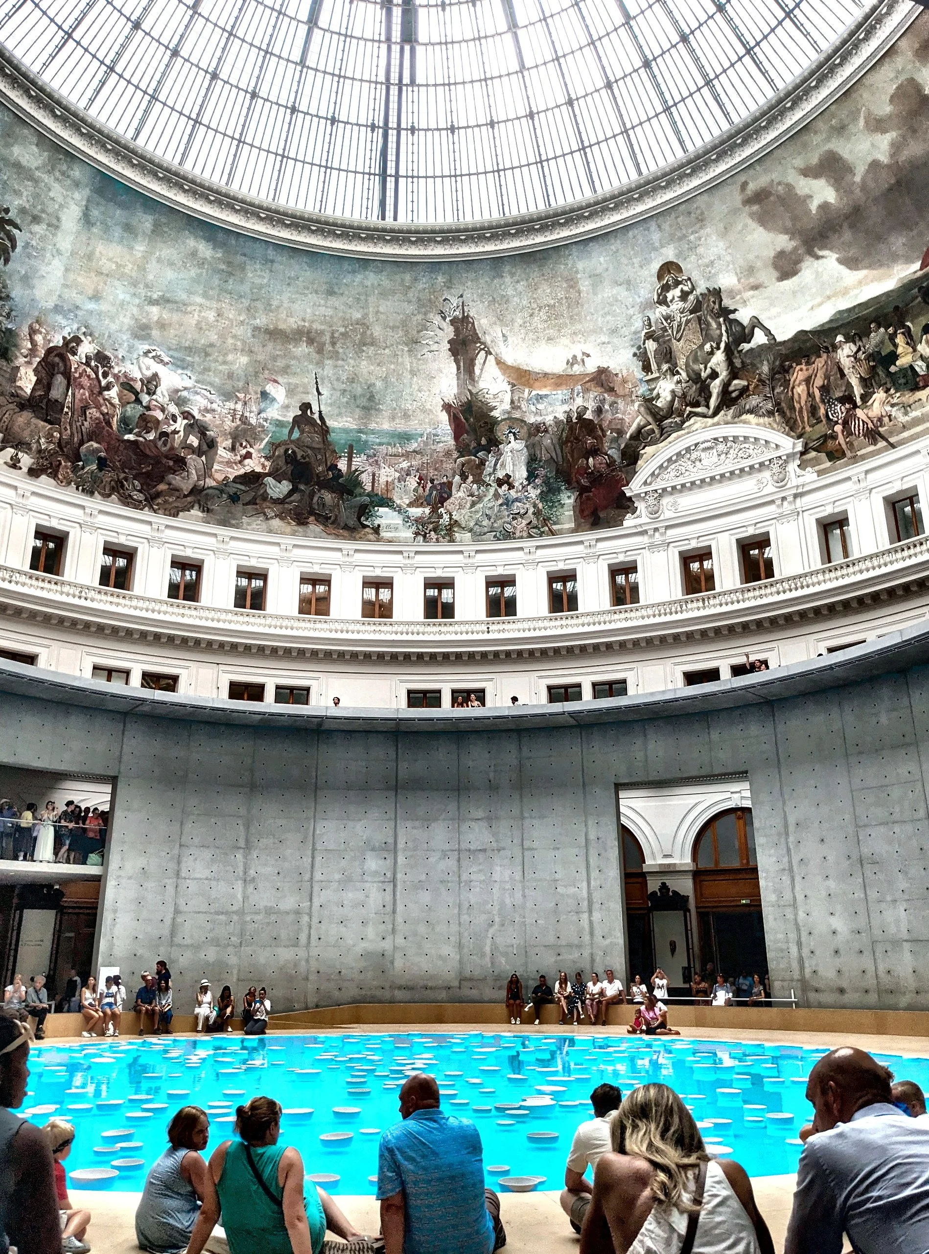 Interior view of a large domed hall with a mural on the upper walls, a pool with floating objects, and people sitting and standing around.