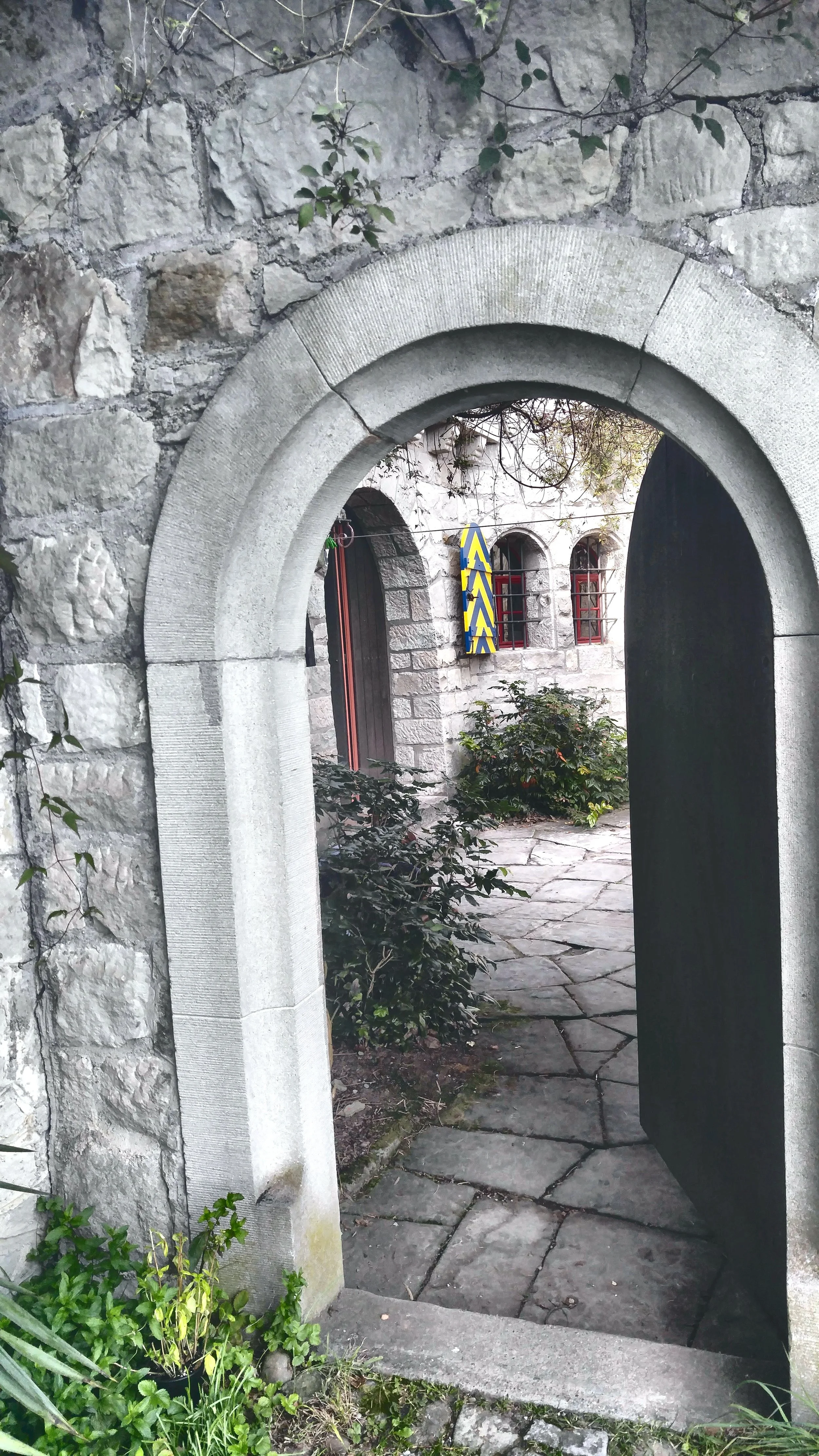 Stone archway leading to a courtyard with stone pathway and plants, view through open black gate.