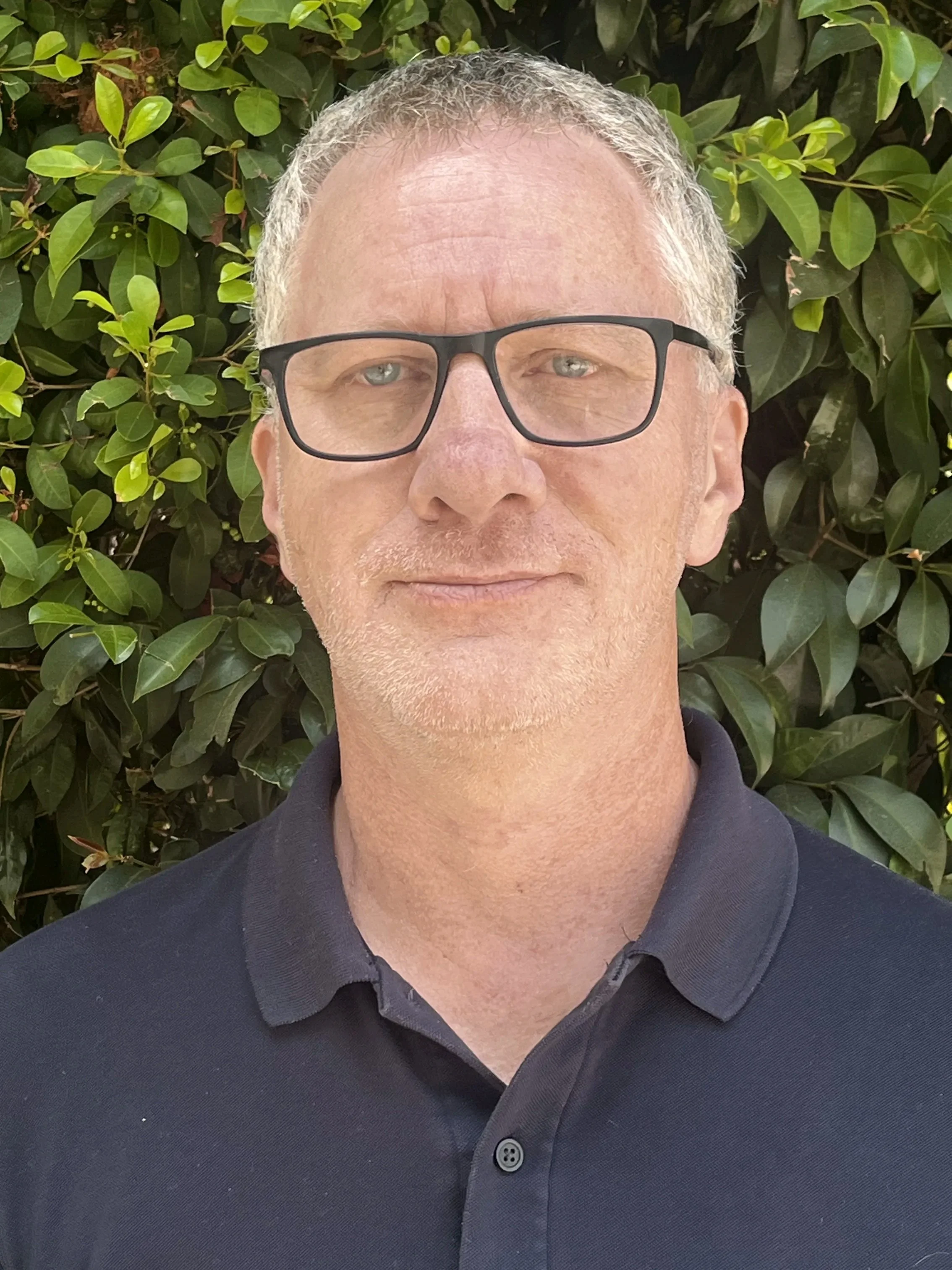 Close-up of a middle-aged man with gray hair, wearing black glasses, in front of green foliage, dressed in a dark collared shirt.
