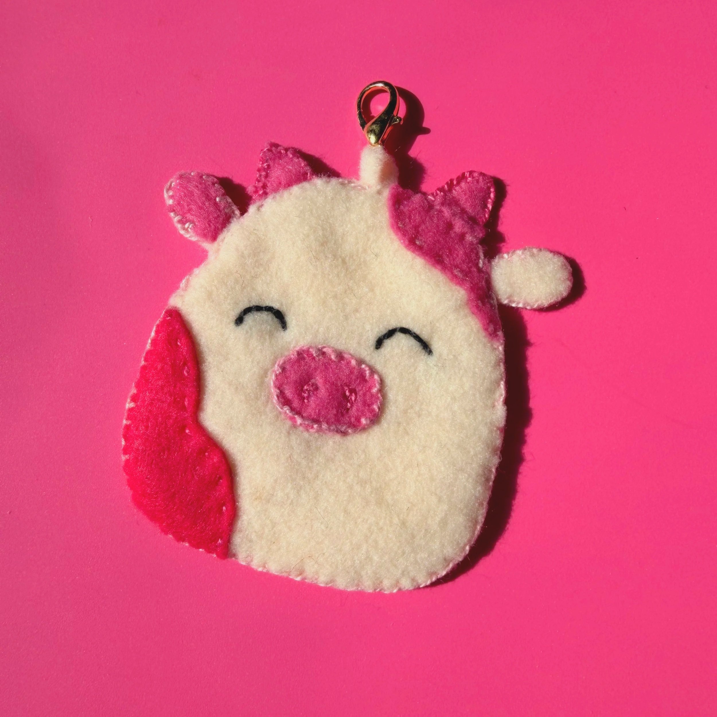 Pink background with a plush unicorn toy featuring pink ears, pink horn, and closed eyes, with a gold earring or accessory attached to the unicorn's head.