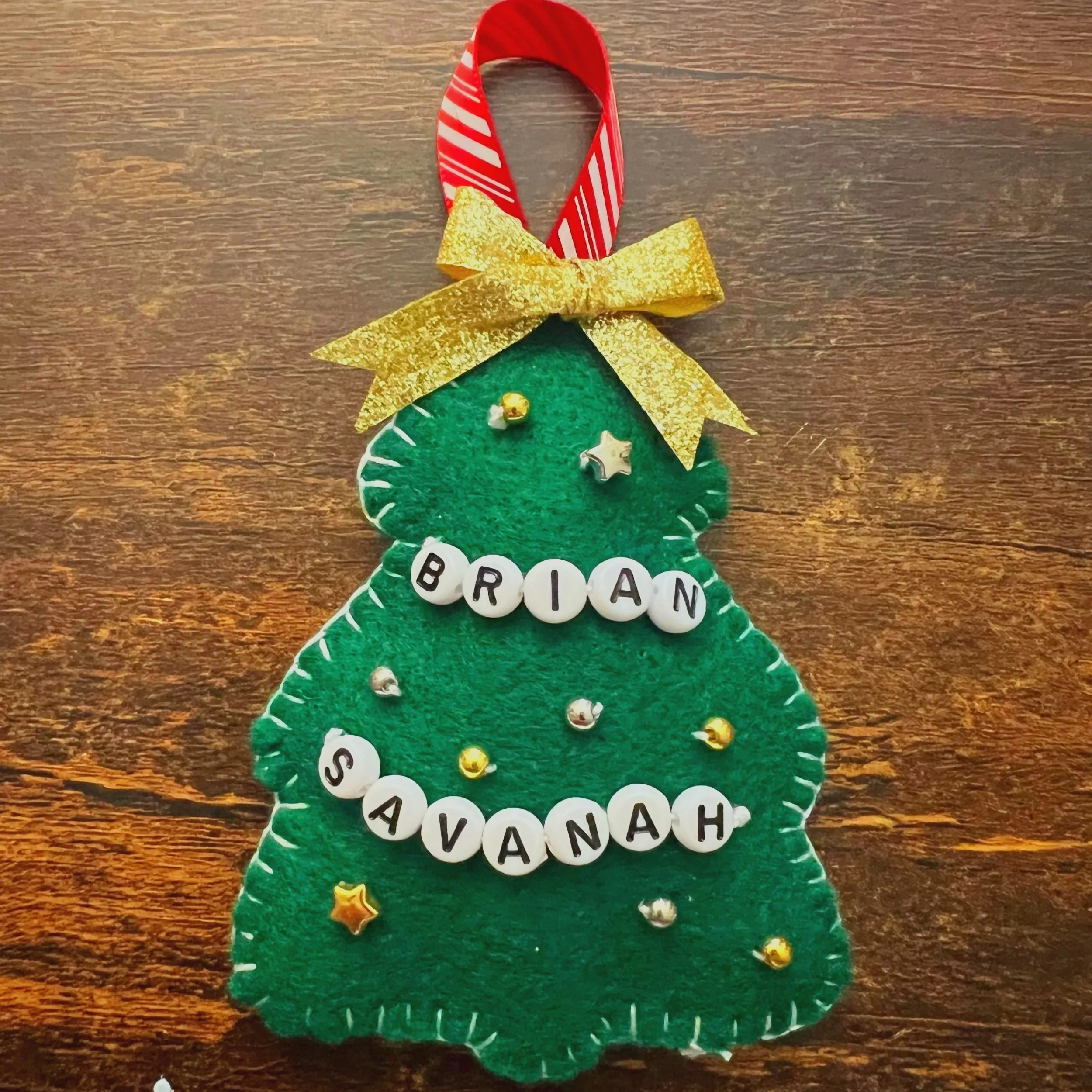 A decorative Christmas tree ornament made of green felt with white stitching, gold, silver, and white stars and round beads, topped with a red and white striped candy cane and a large gold bow.