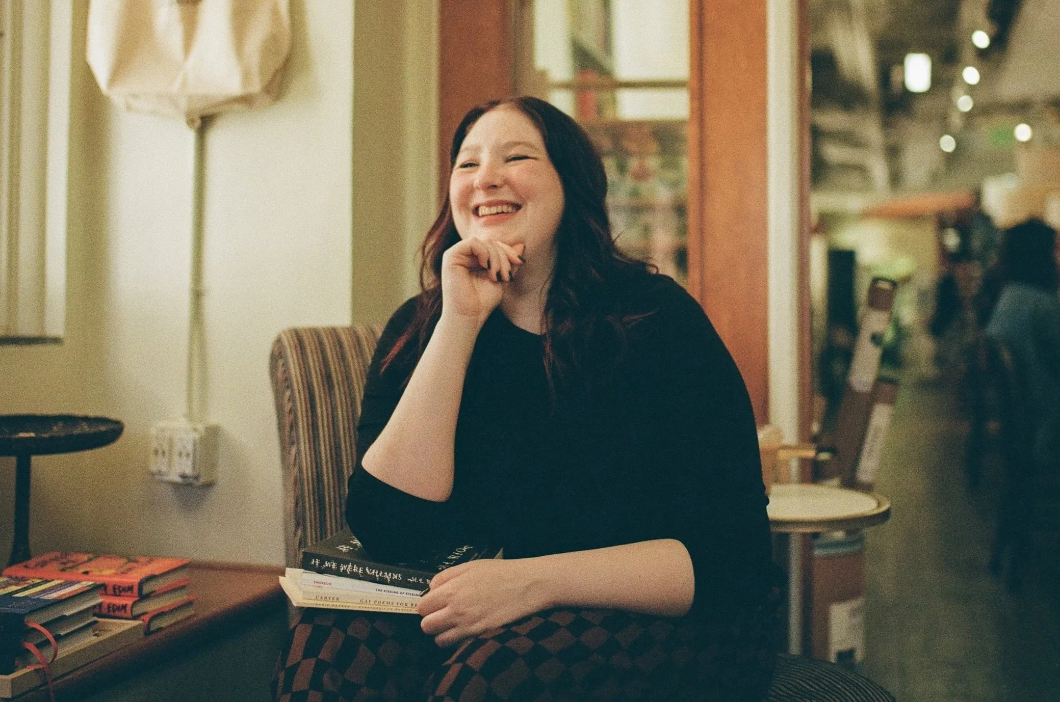A woman with long dark hair smiling while sitting at a table in a cozy indoor space. She is wearing a black top and a checkered skirt, and is resting her chin on her hand. In front of her on the table are several books stacked, and additional books are visible to the side. The background features warm lighting, a stripe-patterned chair, and shelves with various items.