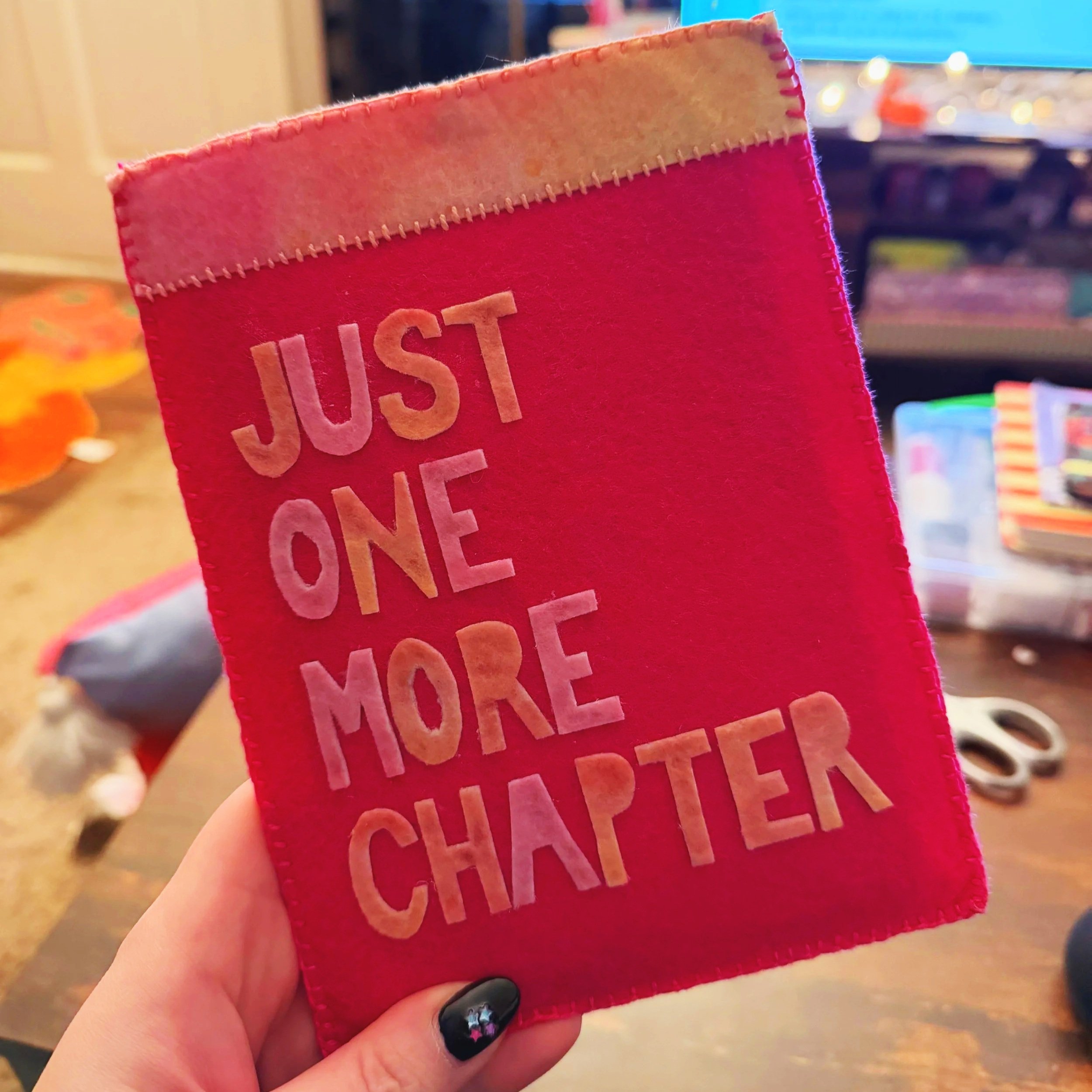 Close-up of a hand holding a small red felt item with beige felt lettering that reads "JUST LOVE" in a blurry indoor setting.