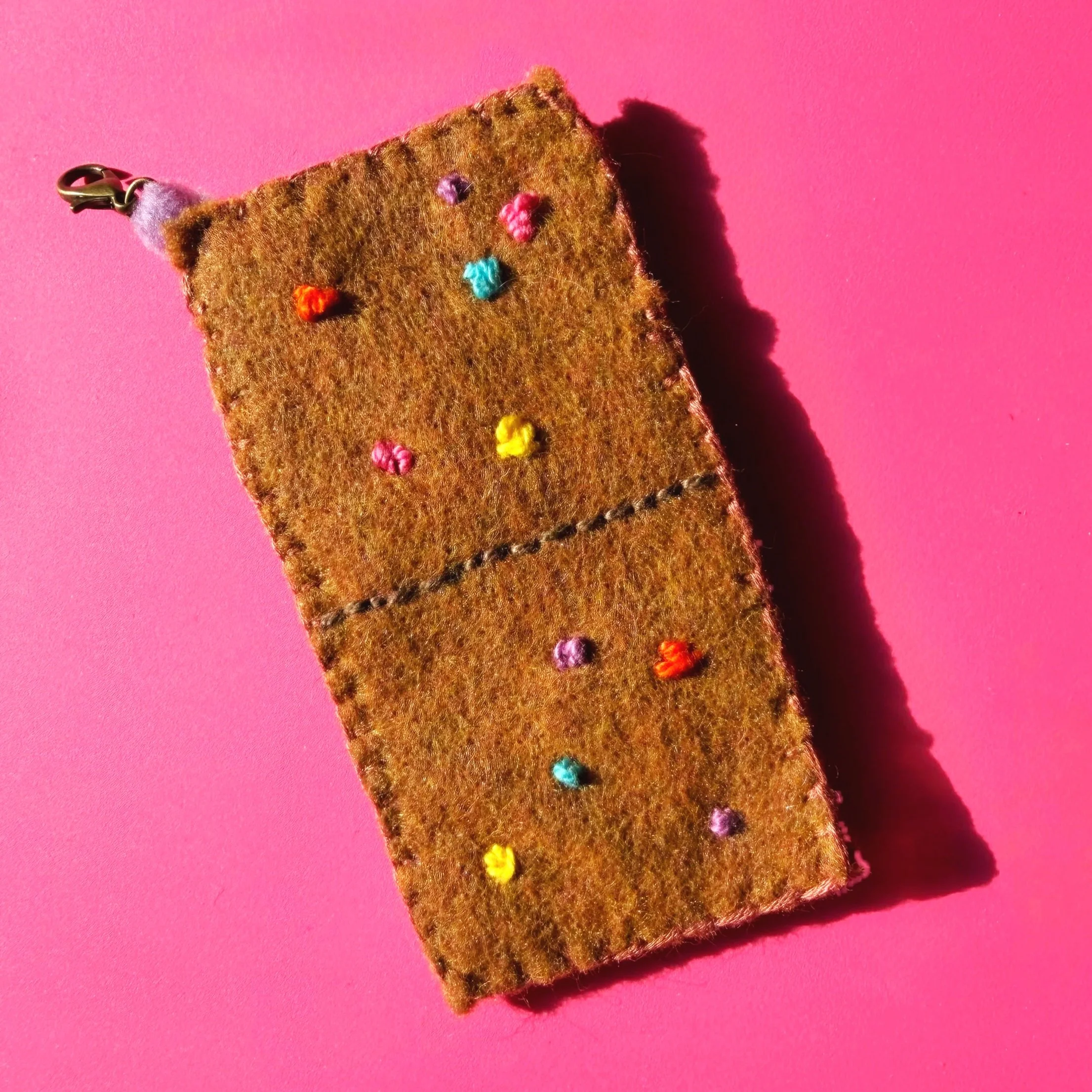Close-up view of a fuzzy, brown, felt-like fabric with multicolored pom-poms, attached to a metal clasp, against a pink background.