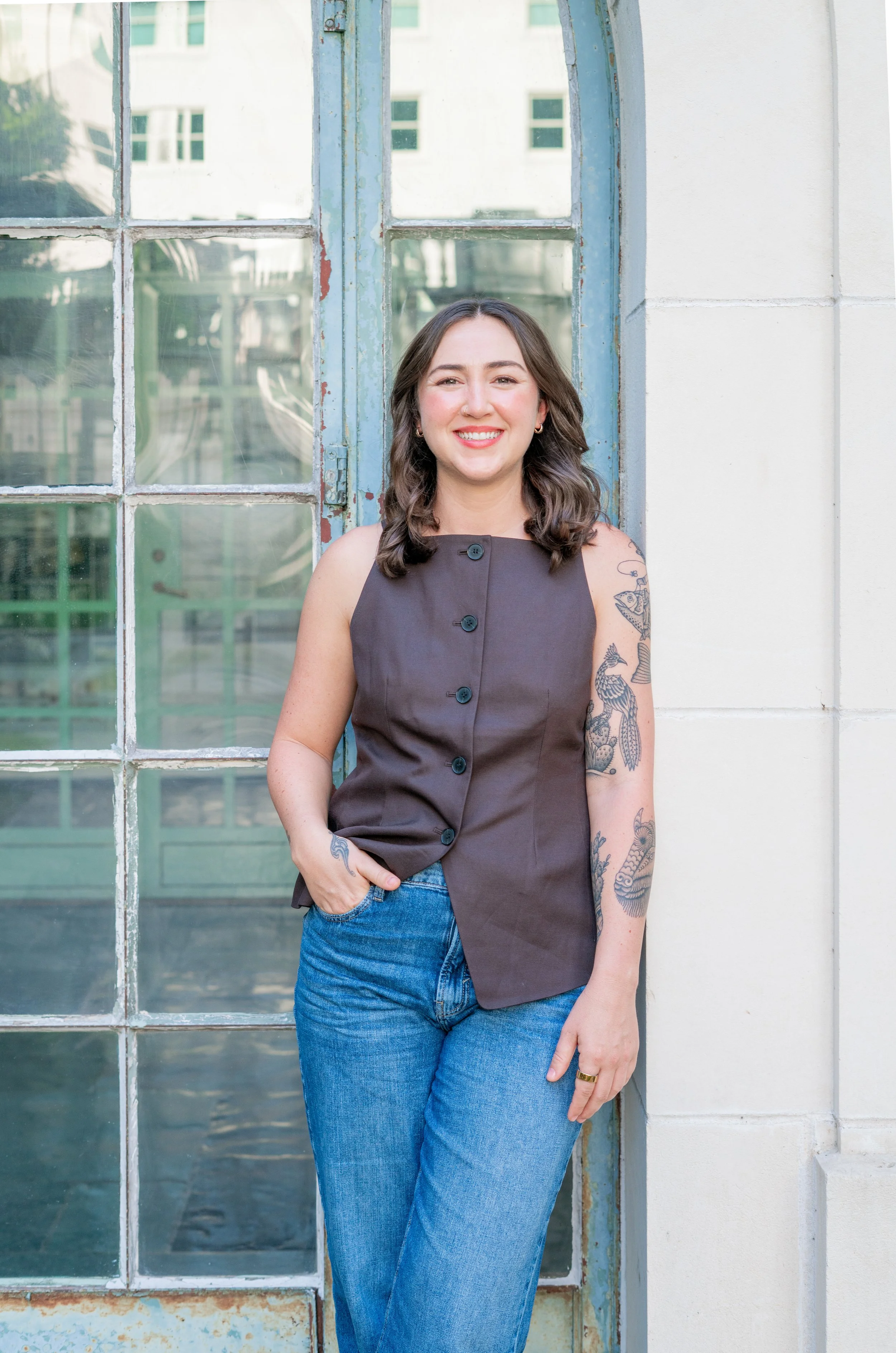 A young woman with dark hair and tattoos on her arm, wearing a sleeveless gray button-up shirt and blue jeans, is standing outdoors in front of an old window with a white wall. She is smiling and has her hand in her pocket.