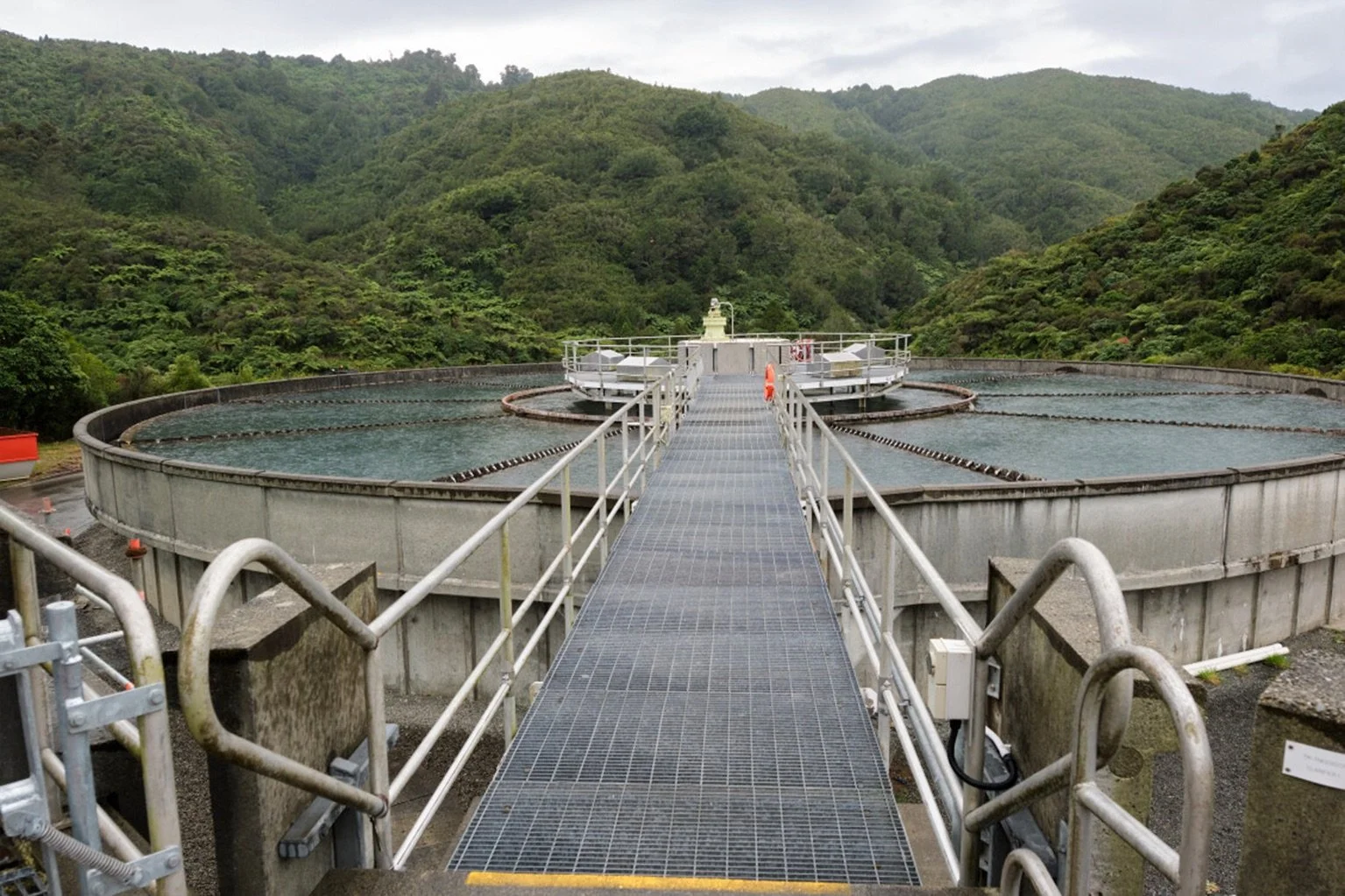 A circular water clarifier at Te Mārua Drinking Water Treatment Plant, with a metal walkway leading to the center, surrounded by green hills.