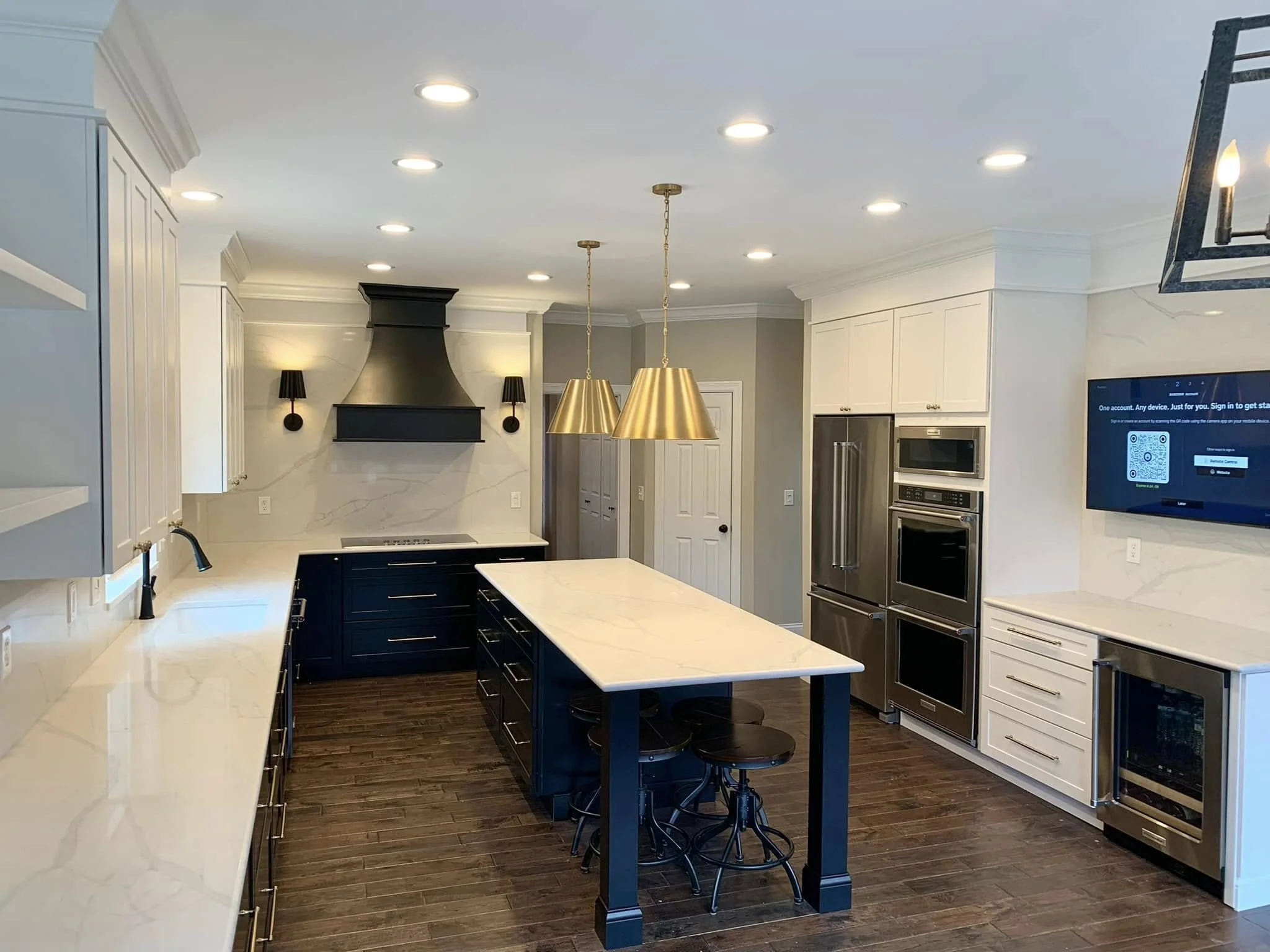 Modern kitchen with white countertops, white cabinets, a black range hood, black and gold pendant lights, a digital screen on the wall, and dark wood flooring.