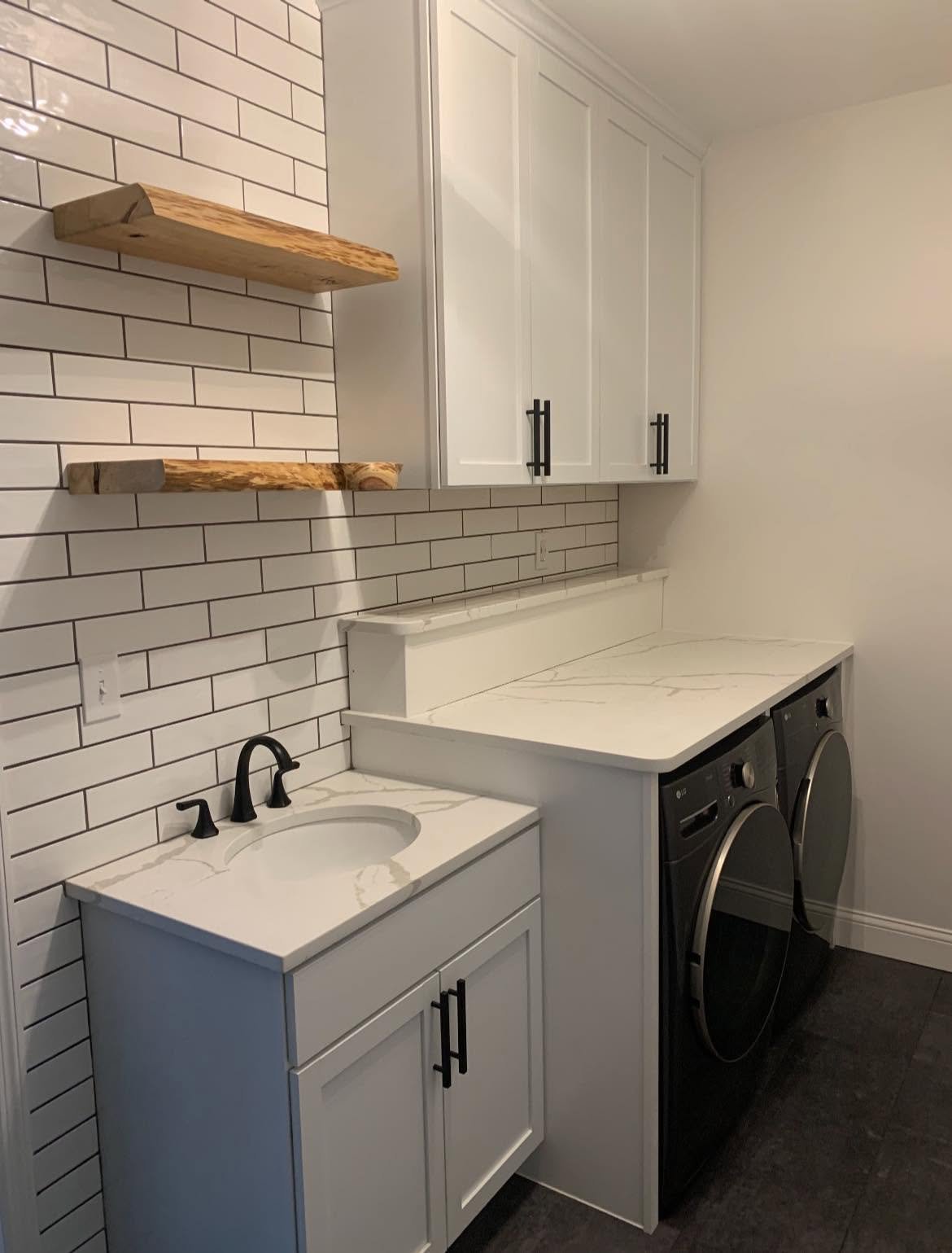 Laundry room with white cabinets, black hardware, a white marble countertop with gray veining, two black front-loading washer and dryer, and a white tiled wall with black grout. Two wooden floating shelves are mounted on the wall.