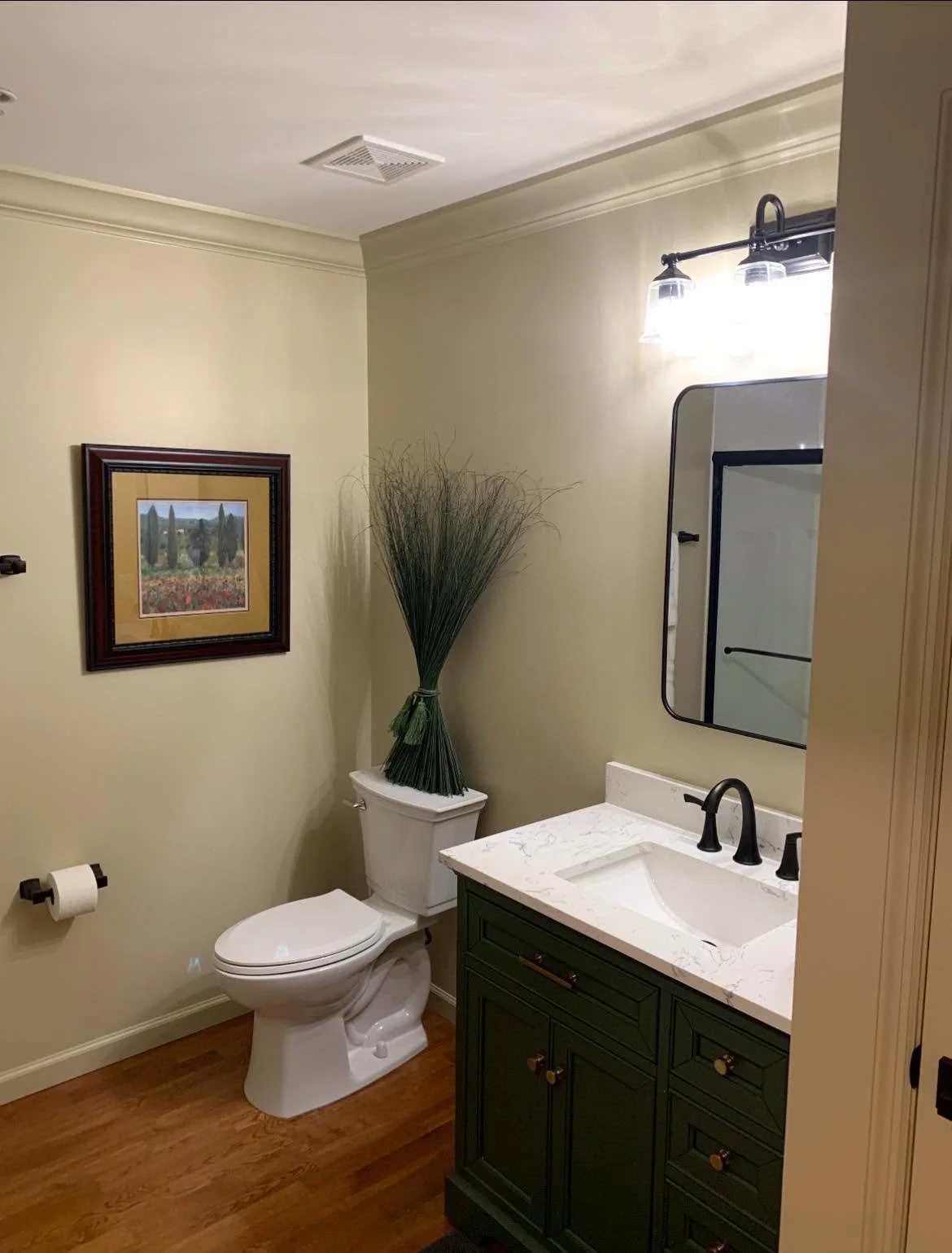 A bathroom with a toilet, a green vanity with a white marble countertop, a rectangular mirror, a wall-mounted light fixture, a framed picture of cypress trees, and a large bundle of decorative grass tied with a green ribbon on top of the toilet tank.