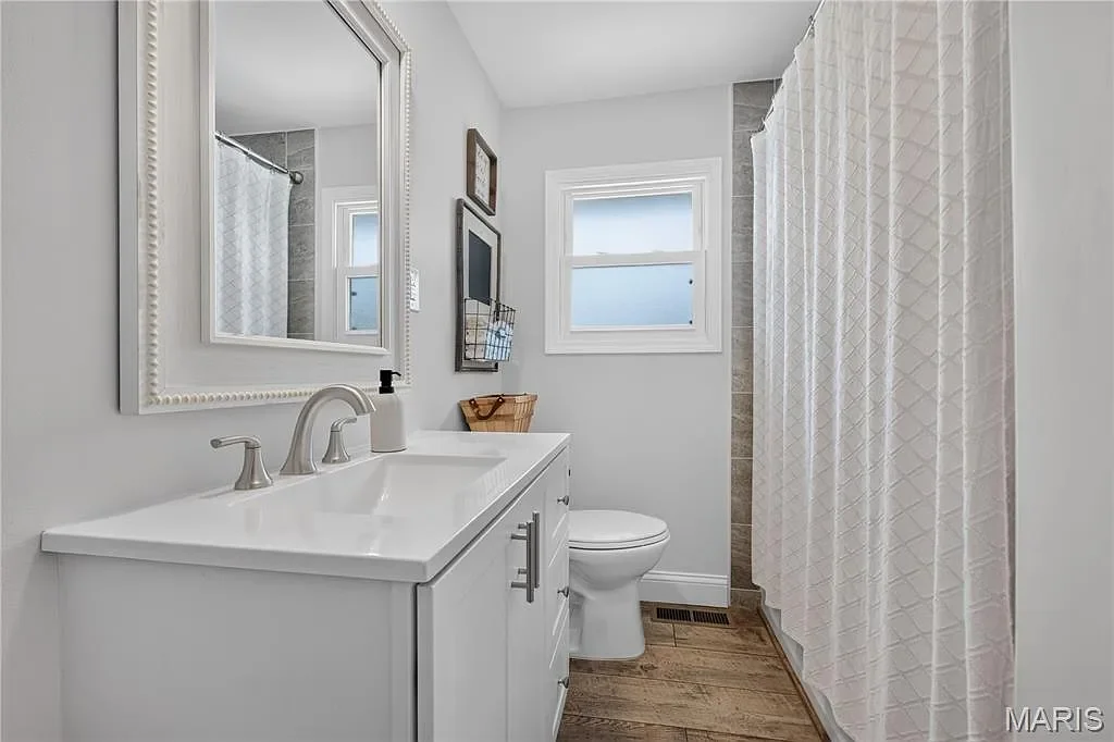 A small bathroom featuring a white vanity with a sink, a mirror, a toilet, and a window with frosted glass. There is a shower with a white curtain and gray tiled walls.