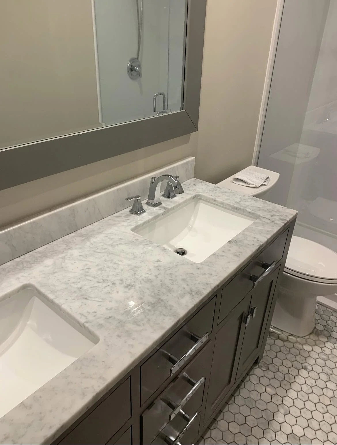 A modern bathroom with a double sink vanity with a marble countertop, a large mirror, a toilet with a folded towel on top, and a glass shower enclosure.