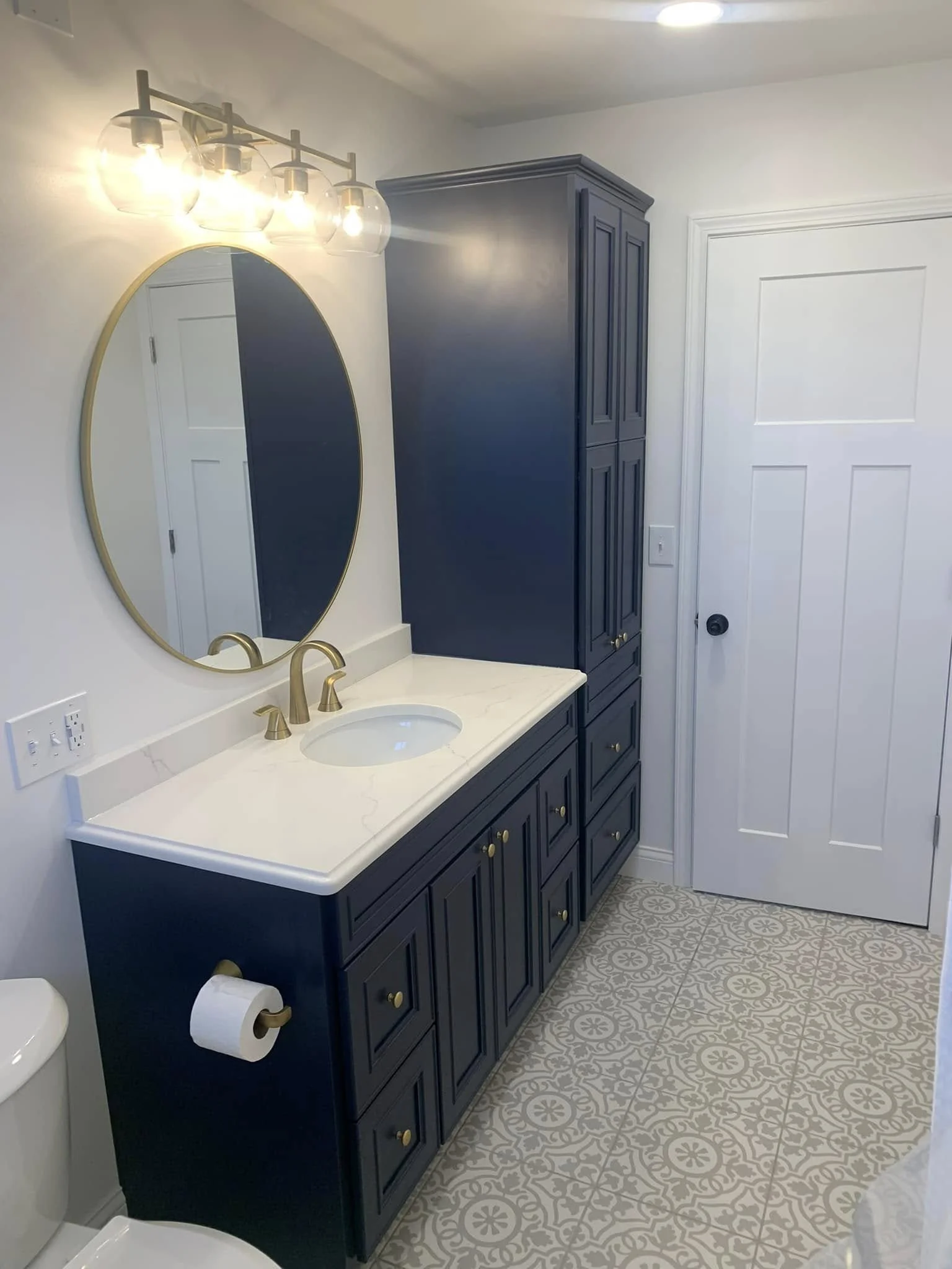 Bathroom with a navy blue vanity, white marble countertop, gold hardware, oval mirror, and patterned tile flooring.