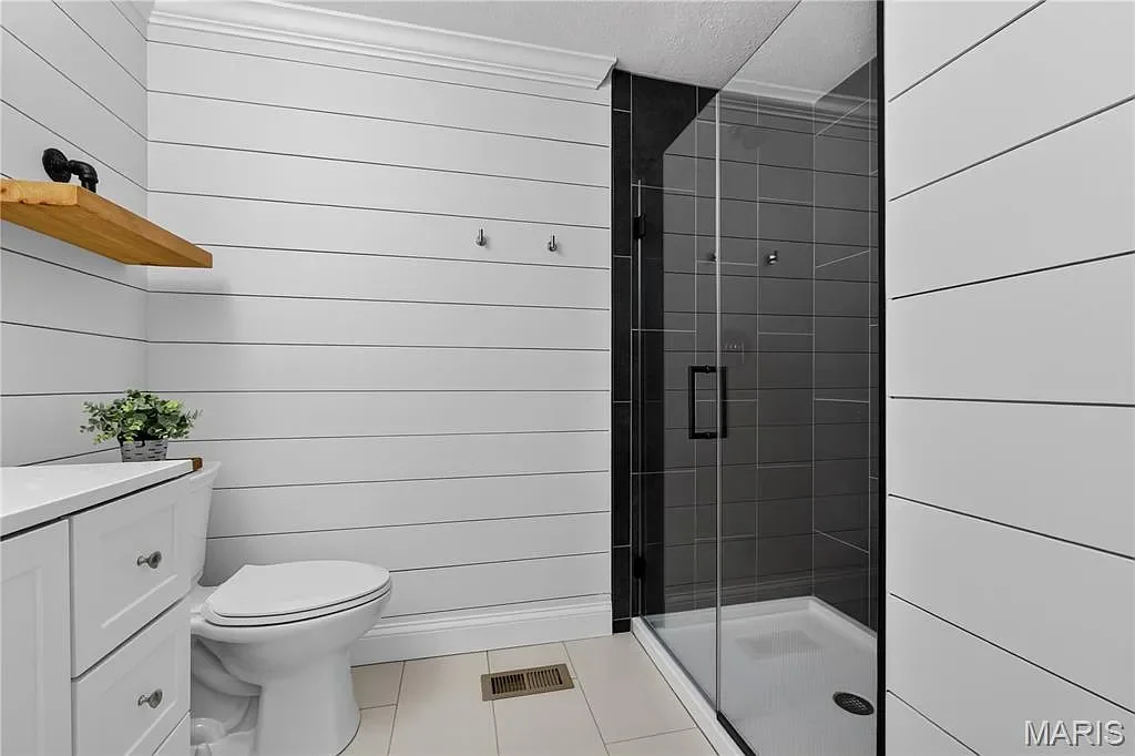 Bathroom with white shiplap walls, a white vanity with a small plant, a toilet, and a glass shower with black tiles and a black door handle.
