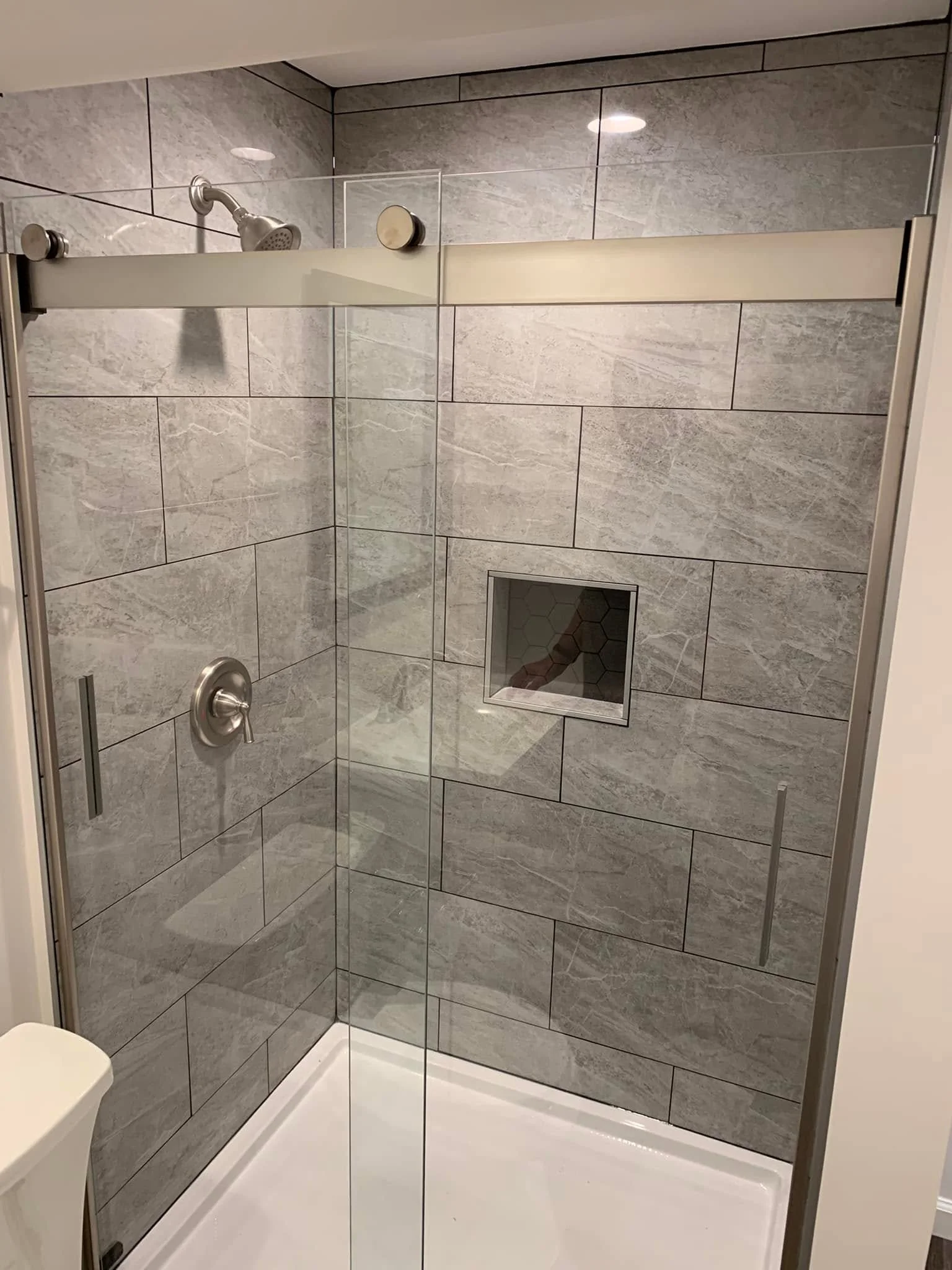 A modern shower with gray tiled walls, a glass door, a showerhead on the left, and a small built-in niche for toiletries.