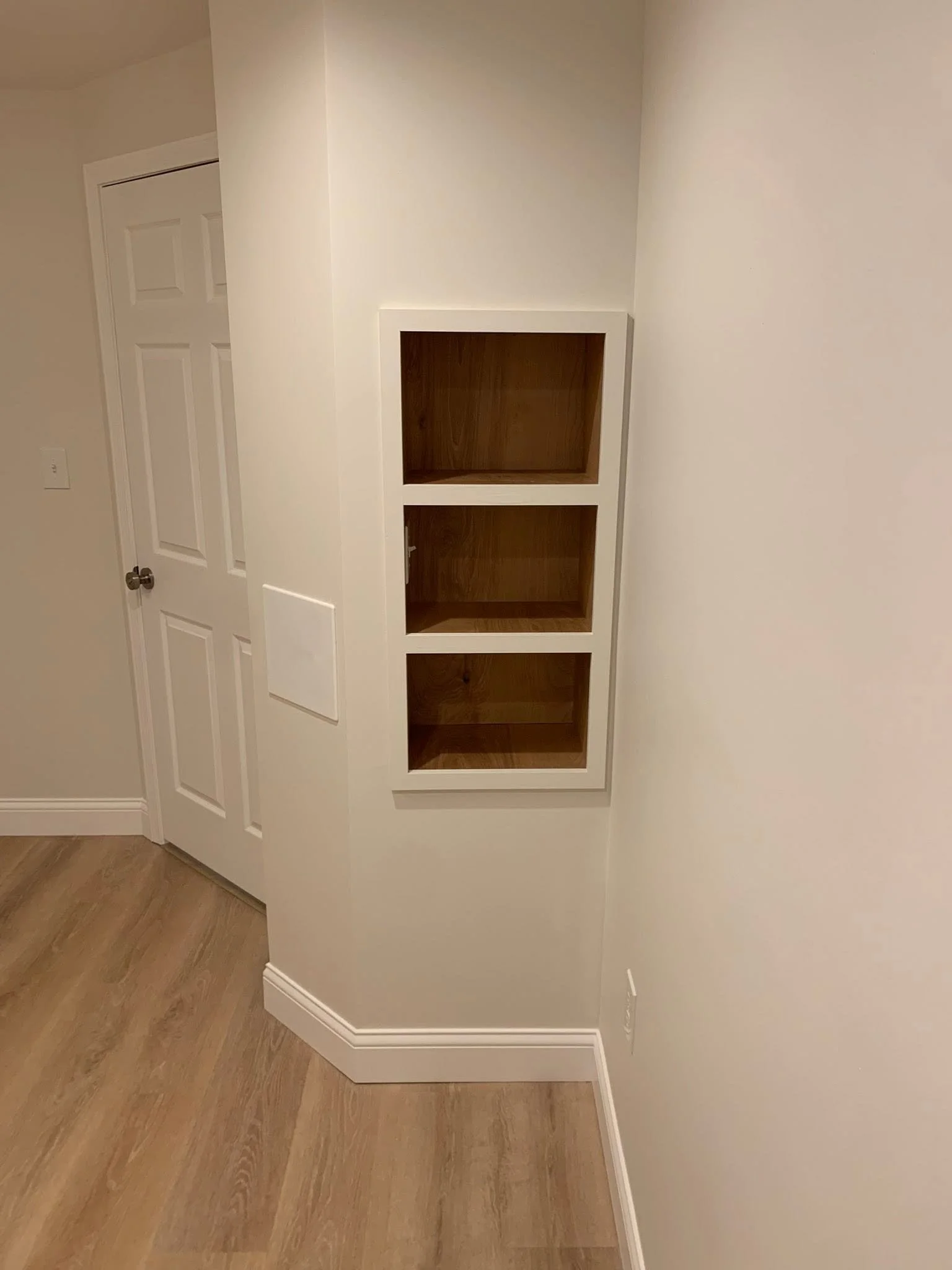 Empty built-in wall shelf with four cubbies in a room with light-colored walls, a closed white door, and wood flooring.