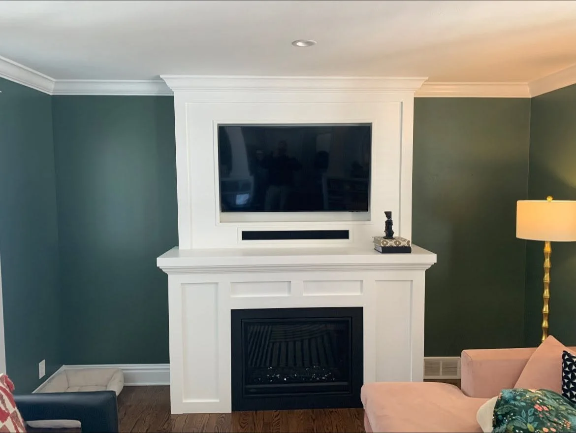 Living room with a white fireplace surround, a mounted flat-screen TV above, a small decorative object on the mantle, green walls, a beige and pink armchair, a tall gold floor lamp with a white shade, and a pet bed on dark wood flooring.