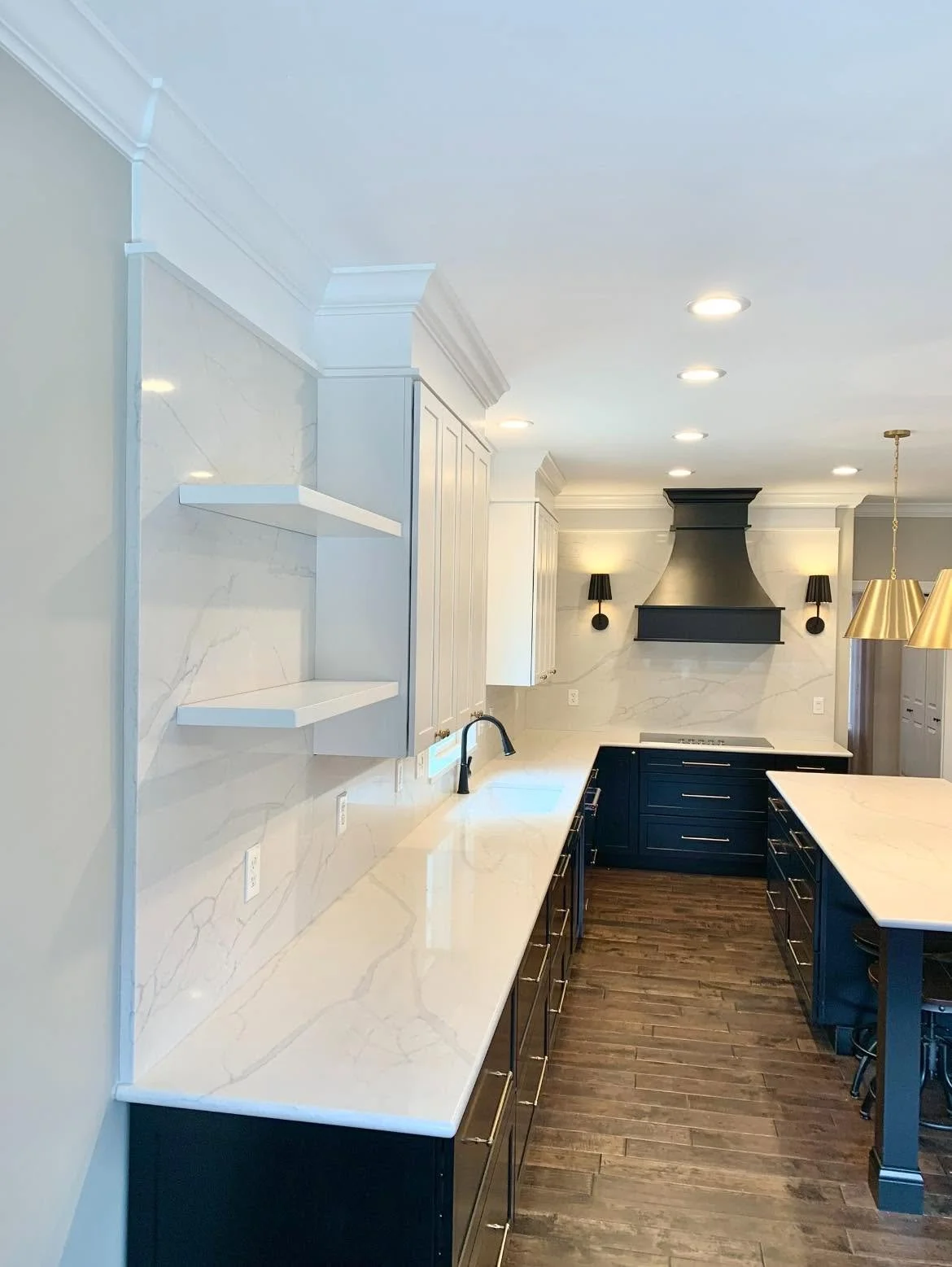Modern kitchen with white marble countertops, dark blue cabinets, white upper cabinets, black vent hood, and black wall sconces, with wooden flooring and a modern gold pendant light.