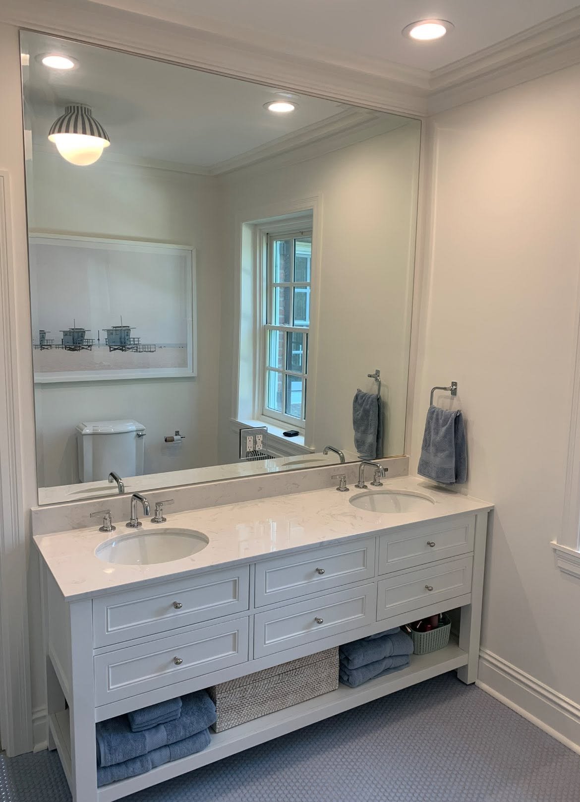 A white double-sink bathroom vanity with folded blue towels and a basket underneath, large mirror above, two gray towels hanging, window with natural light, wall art with lifeguard towers, and ceiling lights.
