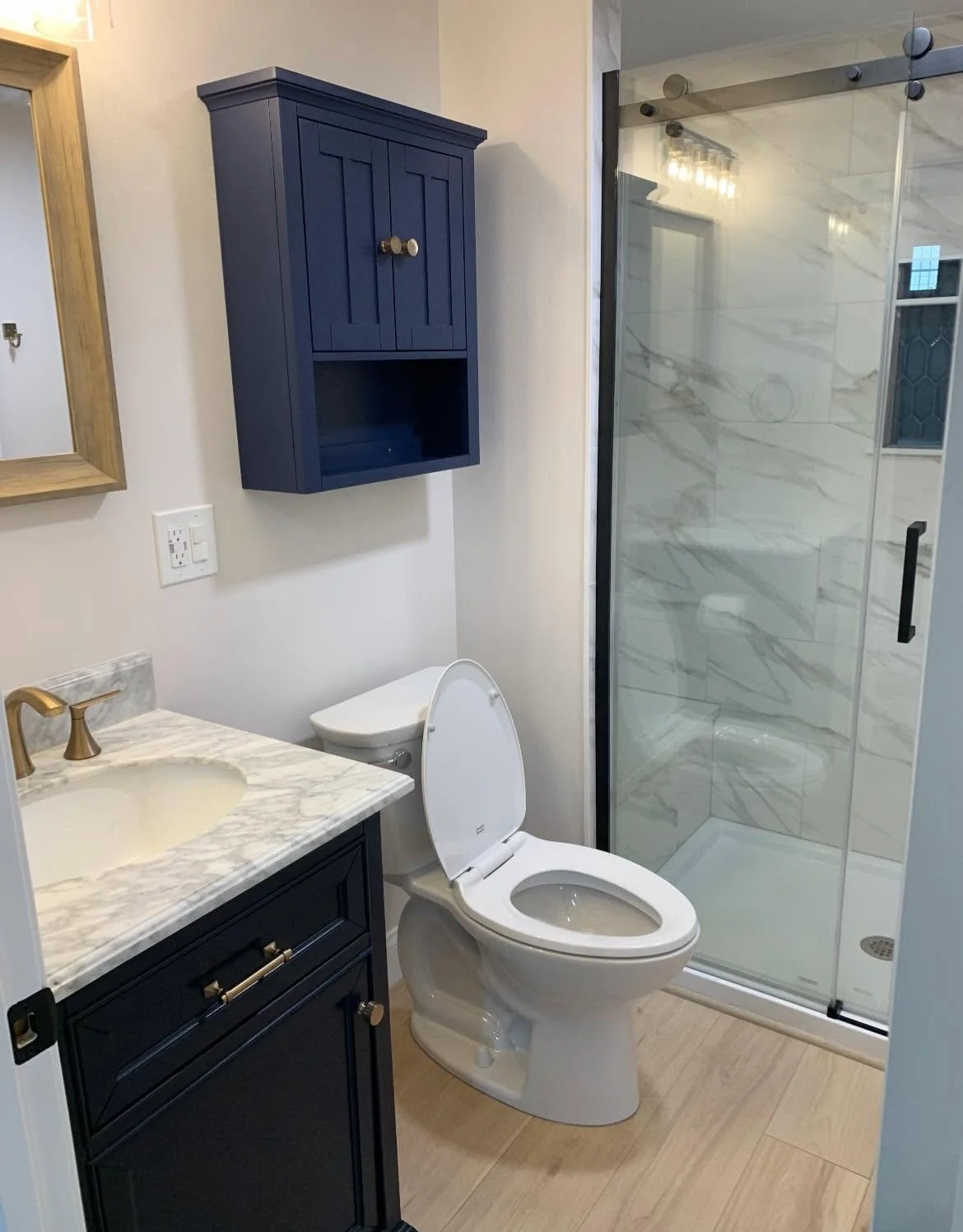A modern bathroom with a white toilet, a navy blue vanity with a marble countertop, a small navy wall cabinet, a glass shower enclosure with marble tile, and a mirror with a wooden frame.