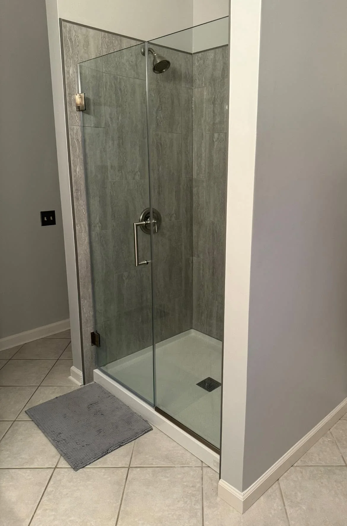 Bathroom shower with glass door, grey tiled walls, silver showerhead, and handle, on beige tiled floor with gray bath mat.
