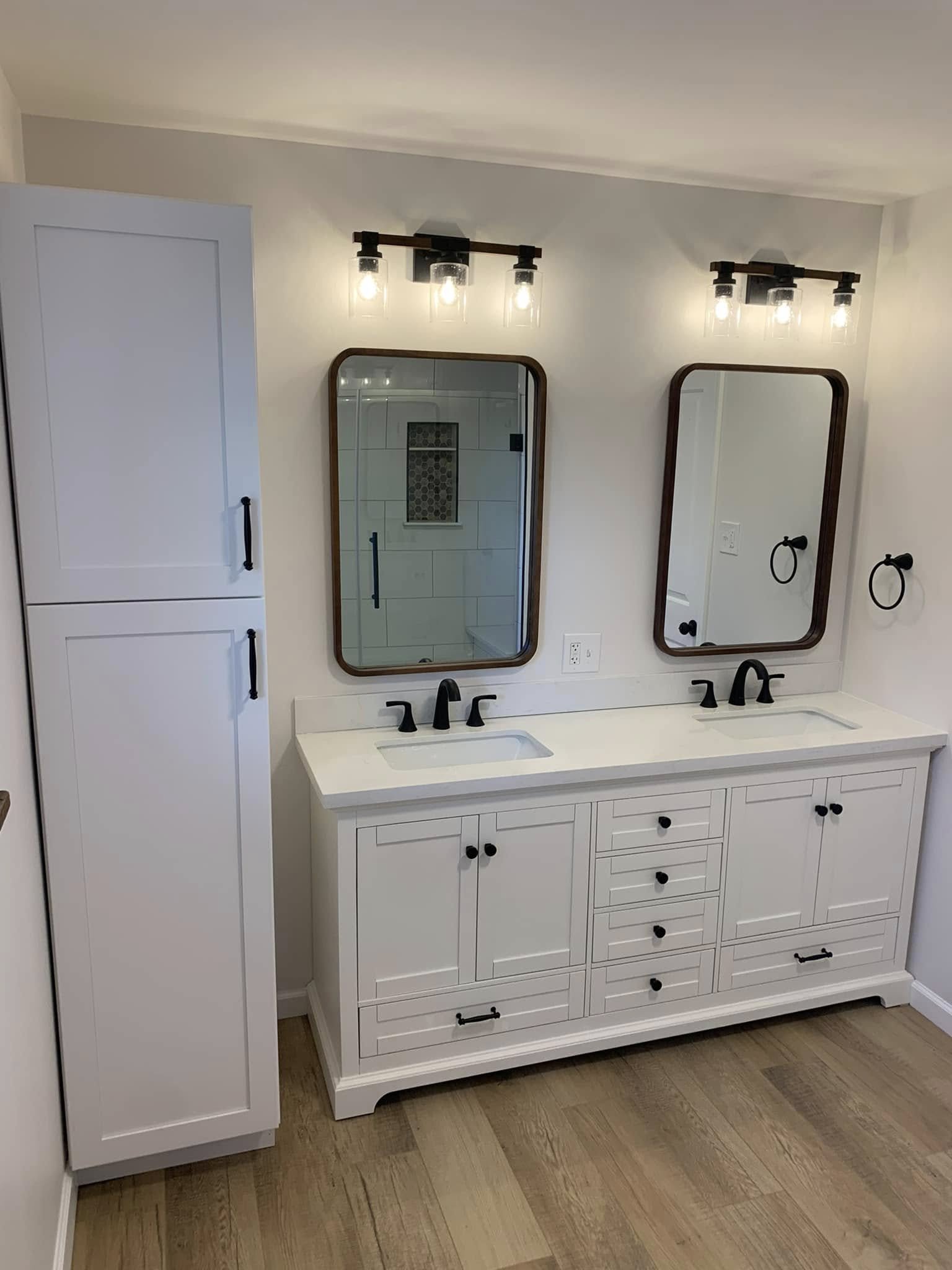 A white bathroom vanity with two sinks, black faucets, and four drawers, topped with two mirrors and two wall-mounted light fixtures with three bulbs each. To the left is a tall white cabinet with black handles, and the background features a shower w