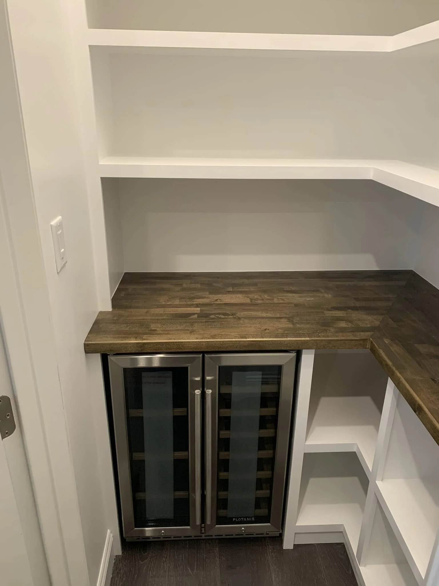 Empty built-in white shelves with a wine refrigerator and wine rack beneath a wooden countertop, situated in a room with dark wooden flooring.
