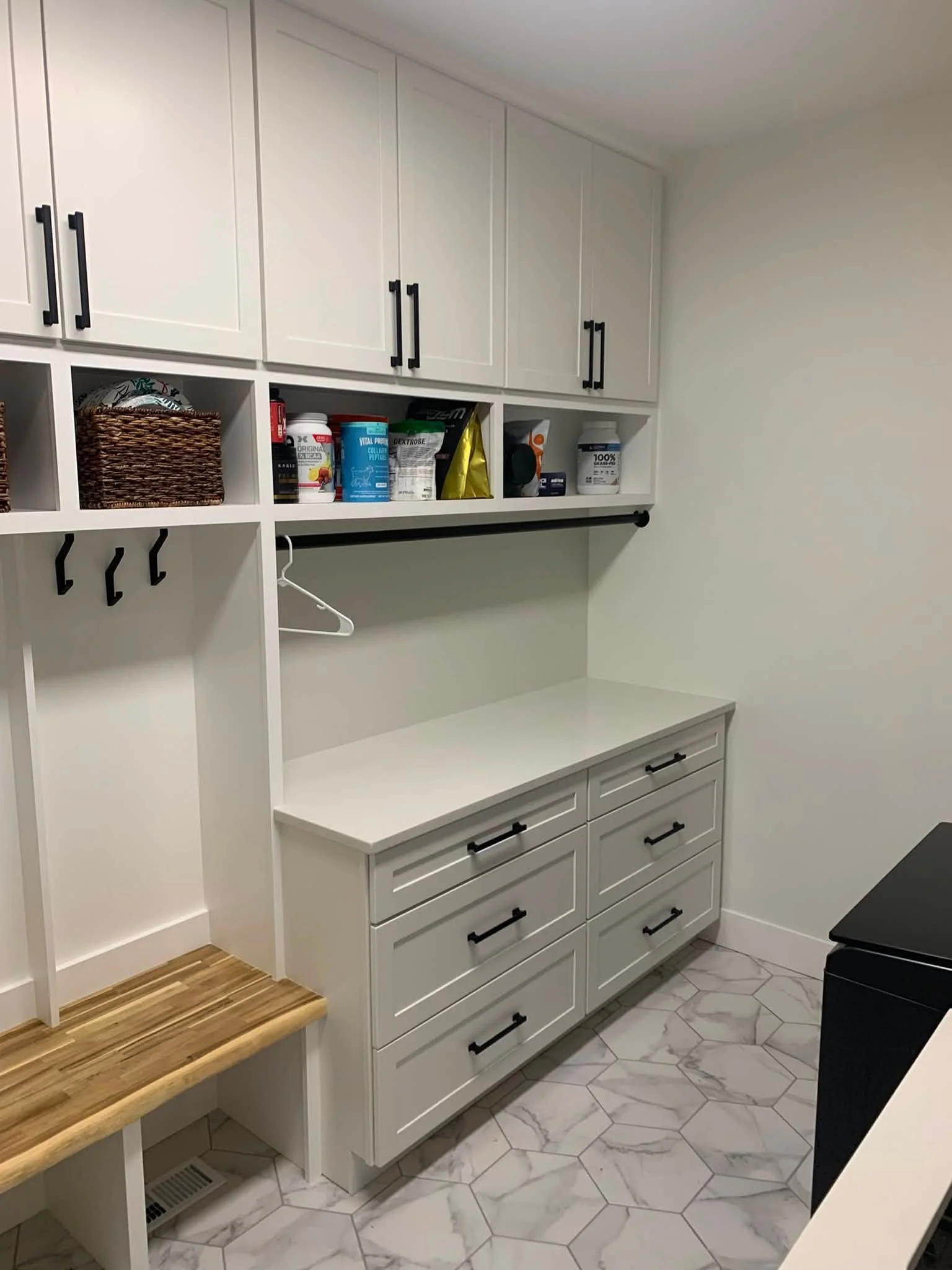 A wall-mounted white cabinet with black handles and drawers, some open cubbies with a woven basket and various supplement containers, a black hanging rod with a plastic hanger, a wooden bench, hexagonal floor tiles, and part of a black appliance.