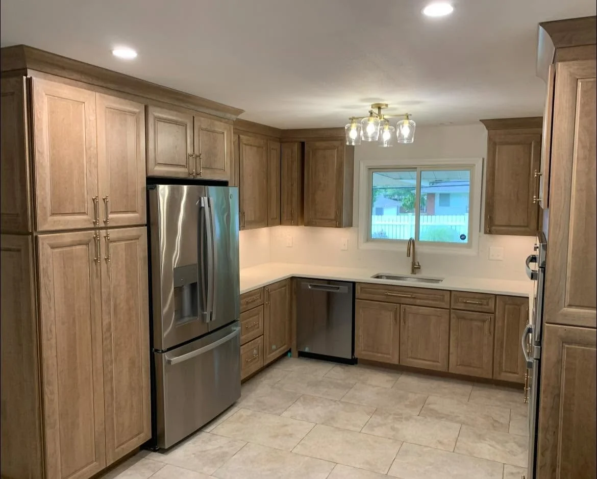 Kitchen with wooden cabinets, stainless steel refrigerator, dishwasher, and a window above the sink, illuminated by ceiling and pendant lighting.