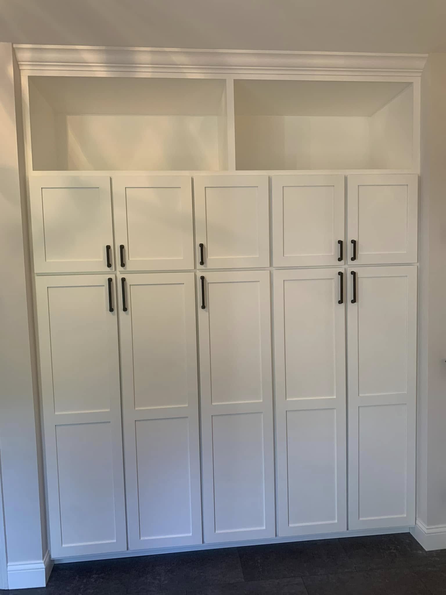 A white built-in cabinet with multiple doors, black handles, and open shelves at the top, located against a white wall.