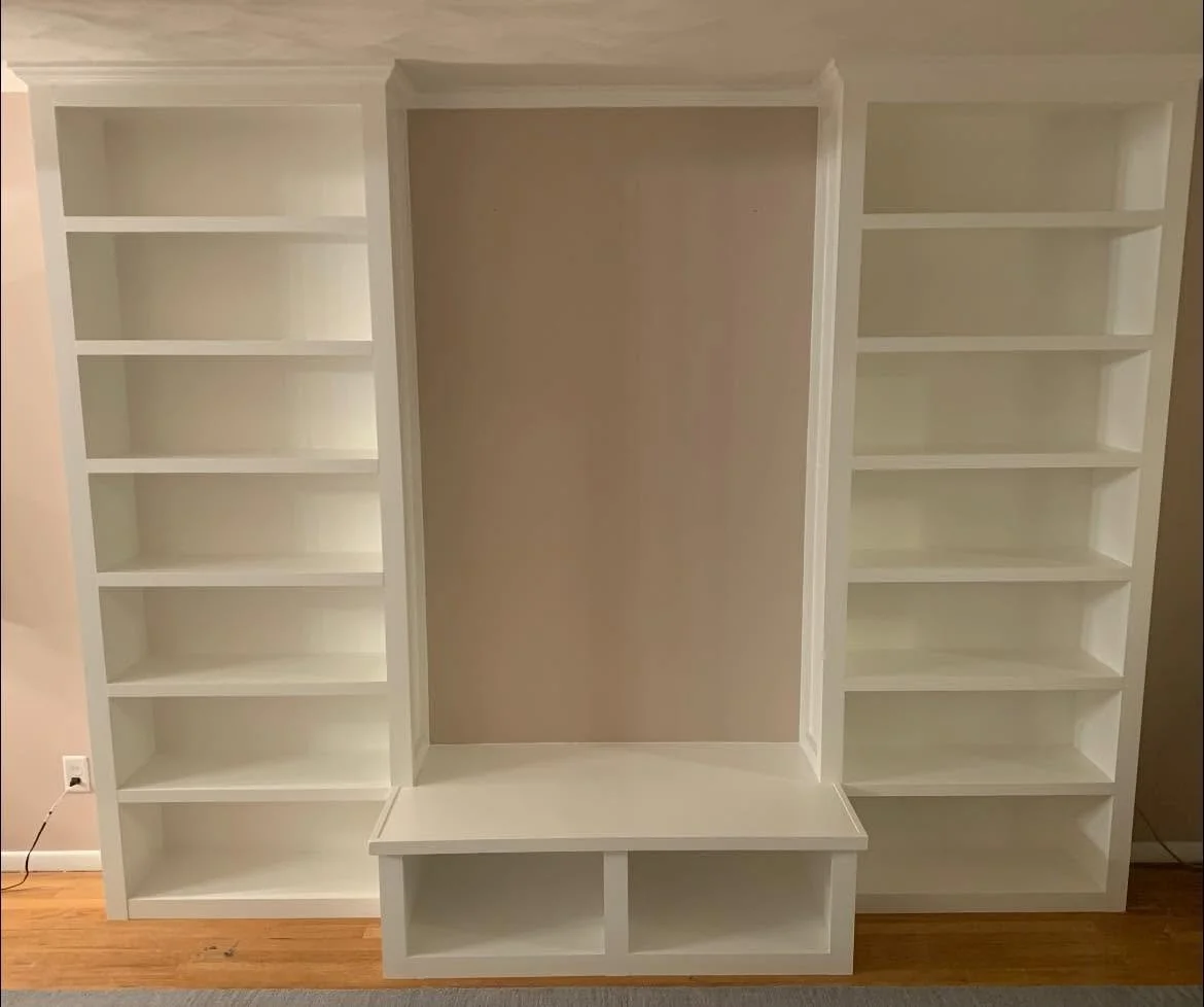 Empty built-in white bookshelf with a central niche and a bench with storage compartments at the bottom, against a beige wall.